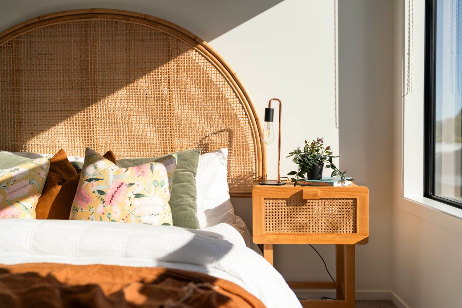 Sunlit modern bedroom interior with rattan headboard, wooden nightstand and white bedding in warm morning light.