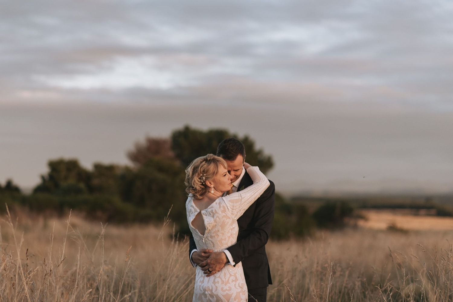 A romantic couple embraces in a golden field at sunset as dramatic clouds fill the dusky sky above them.