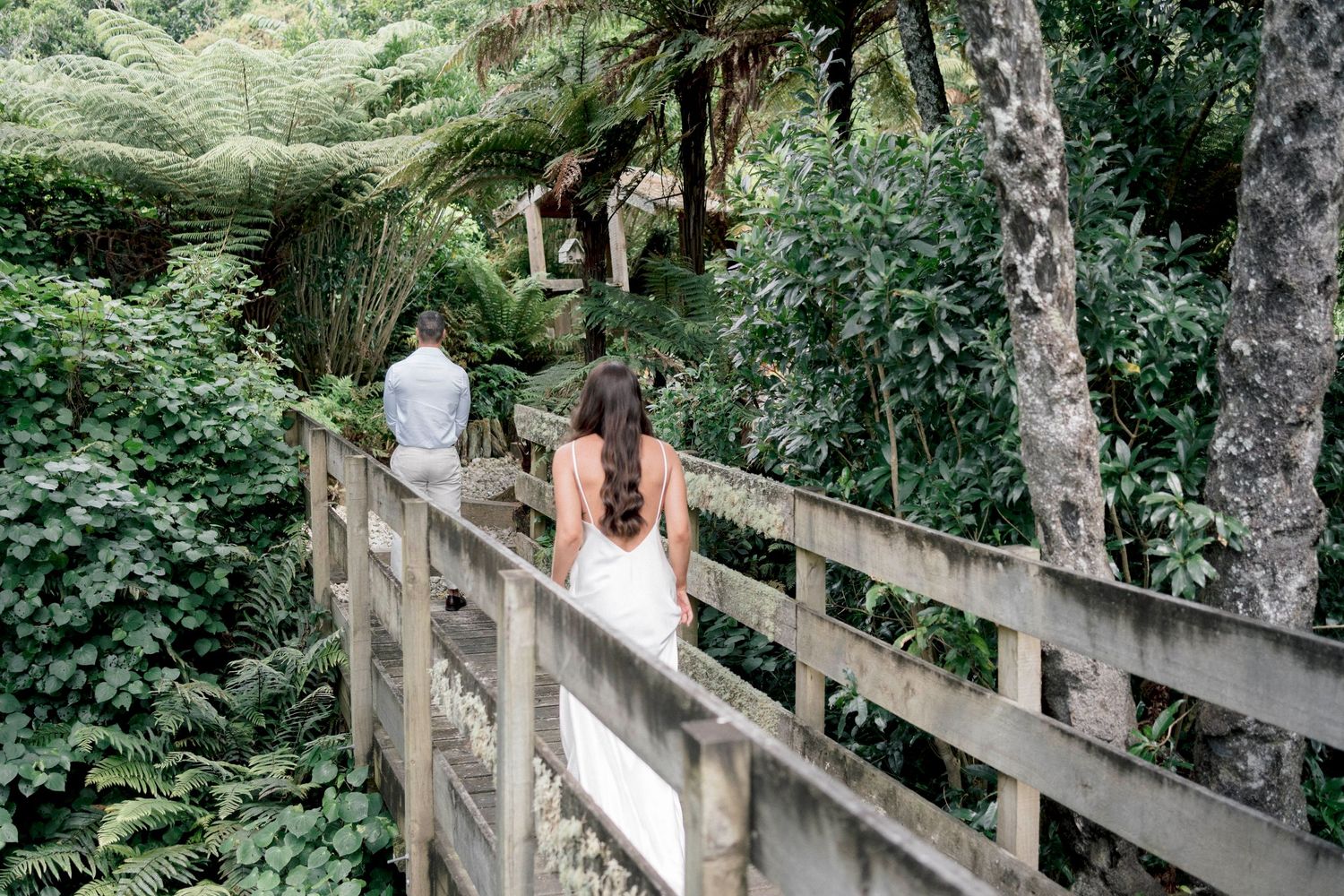 Two people stand on a wooden footbridge surrounded by lush green forest foliage.