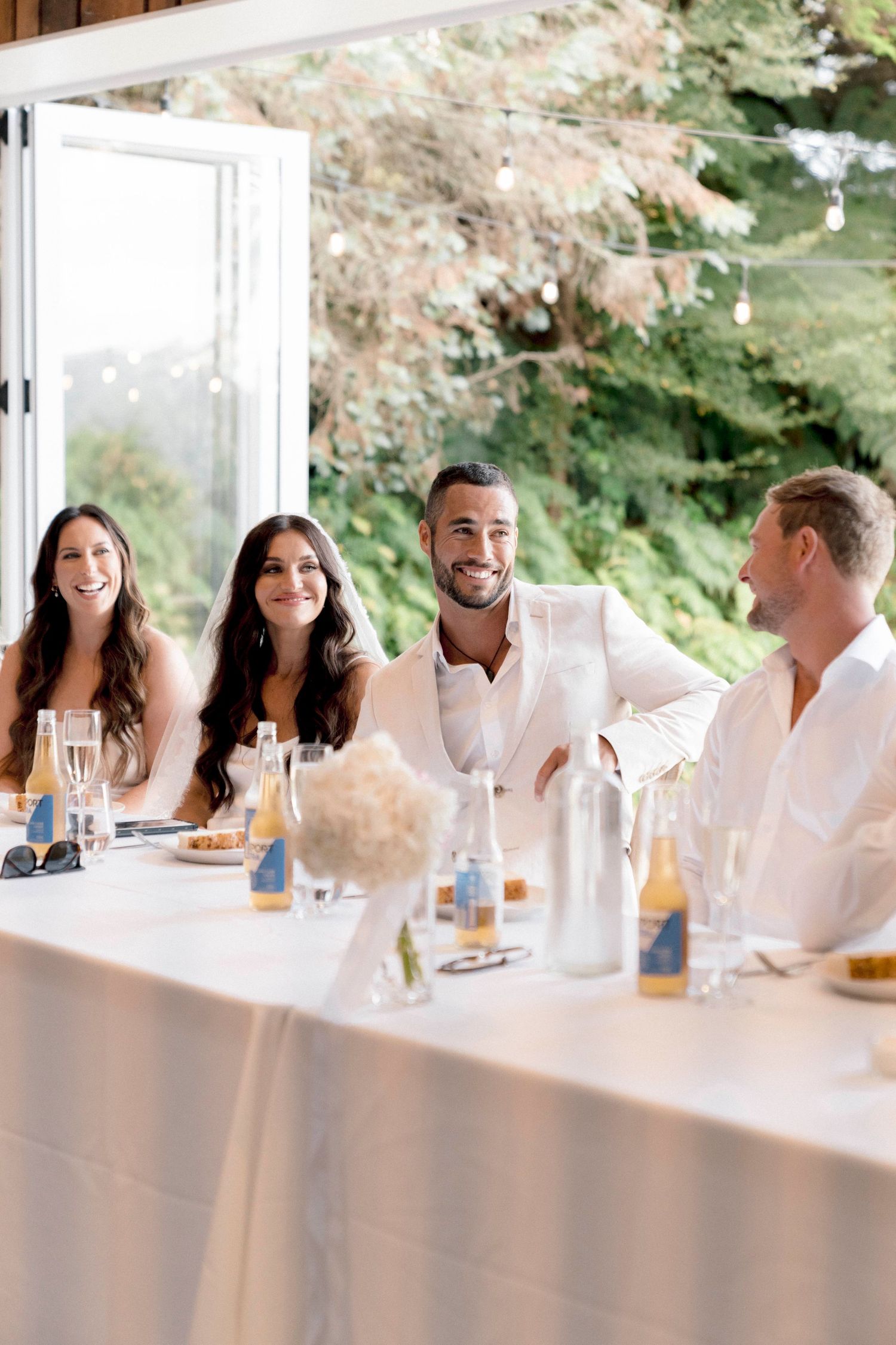 Wedding party shares laughs at elegant white reception table setting.