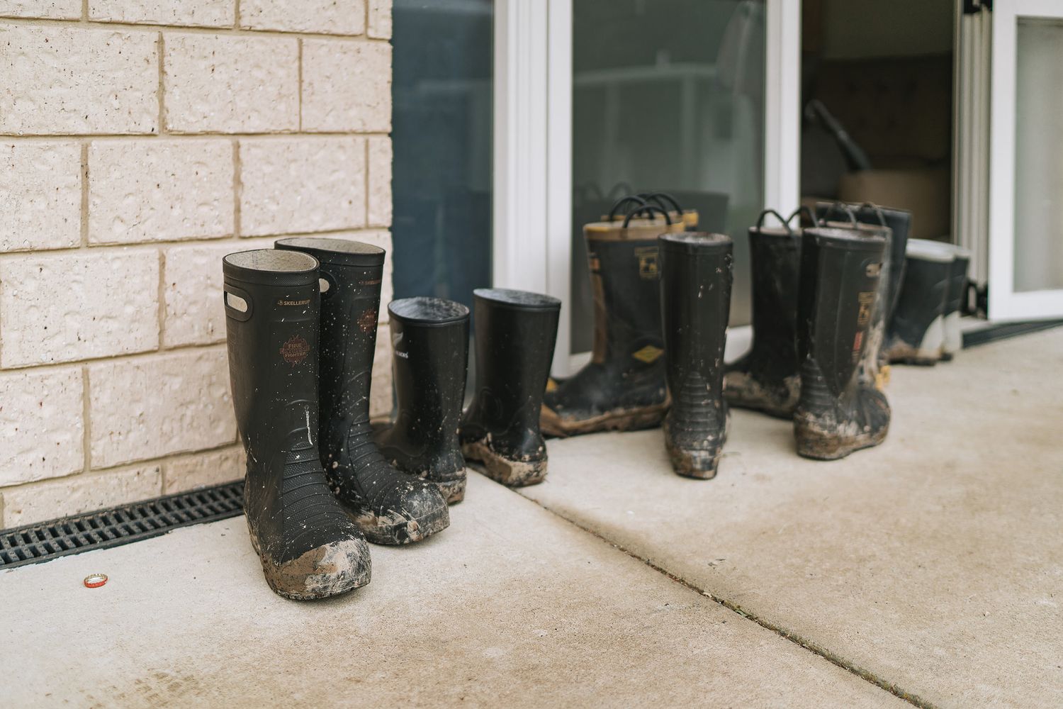 A row of black rubber rain boots lined up on a beige concrete porch against a white brick wall.