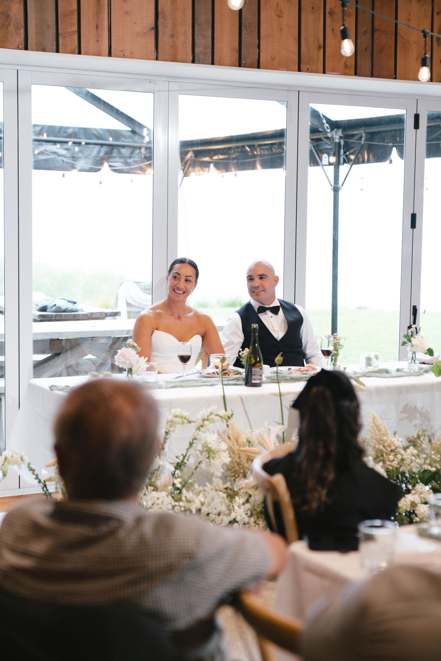 Wedding guests seated at long tables during an indoor reception with large windows and natural light.