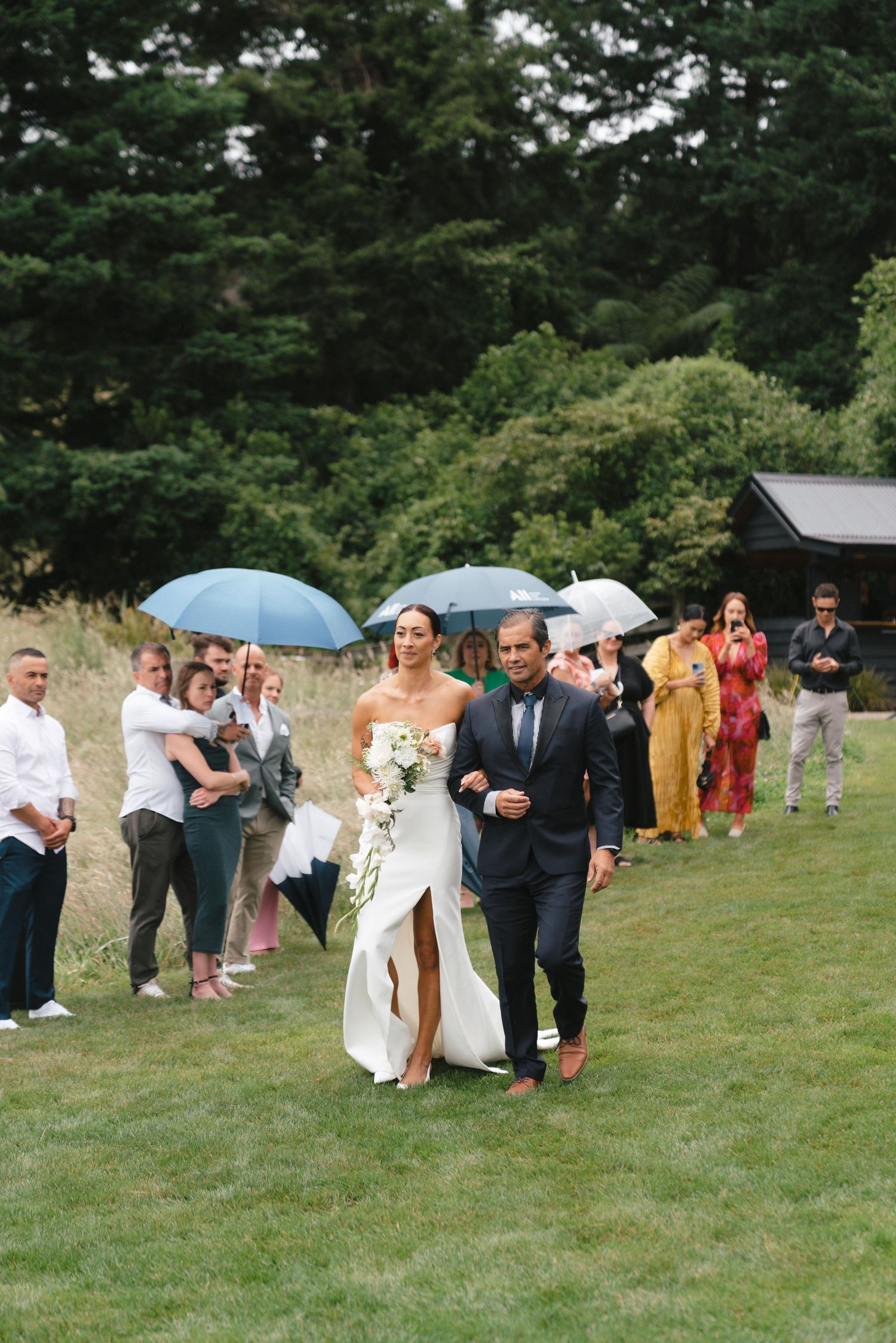 Wedding ceremony held outdoors with guests holding umbrellas on a grassy lawn surrounded by evergreen trees.