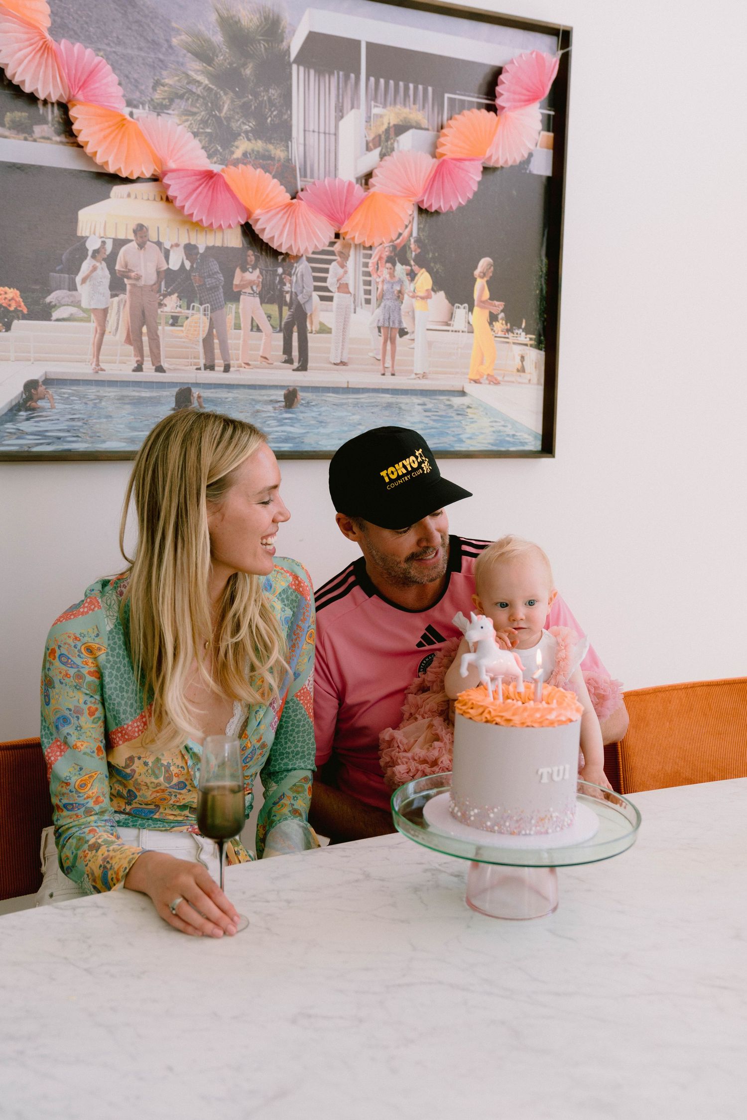 Family celebrates a baby's birthday with pink decorations and cake at a party with coral paper fans hanging above.