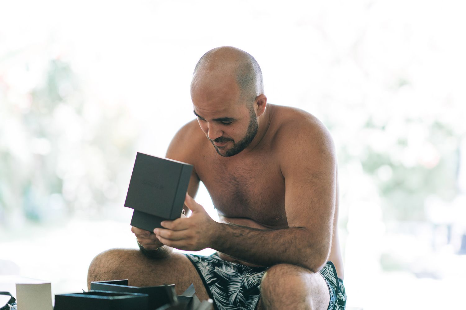 A shirtless man reads intently from a small book while sitting in a bright, airy space.