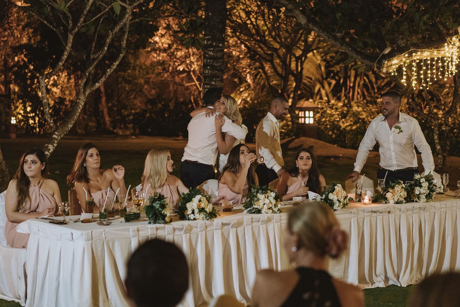 Wedding party members seated at a long white-draped head table during an outdoor evening reception.