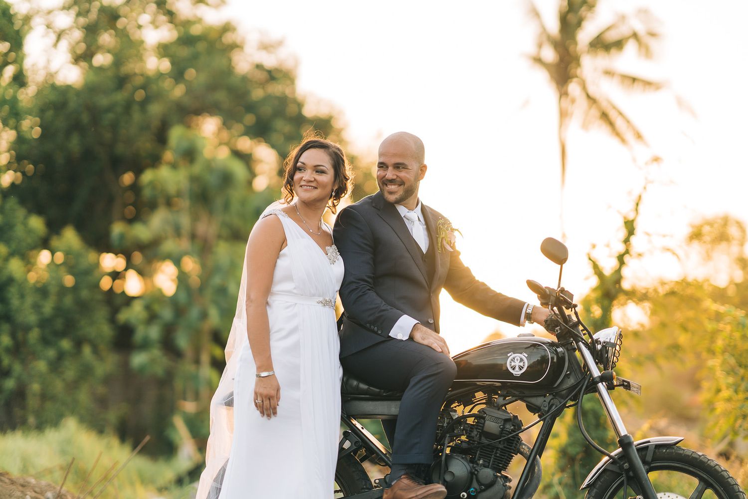 Wedding couple poses together on a vintage motorcycle during golden hour with palm trees in background.