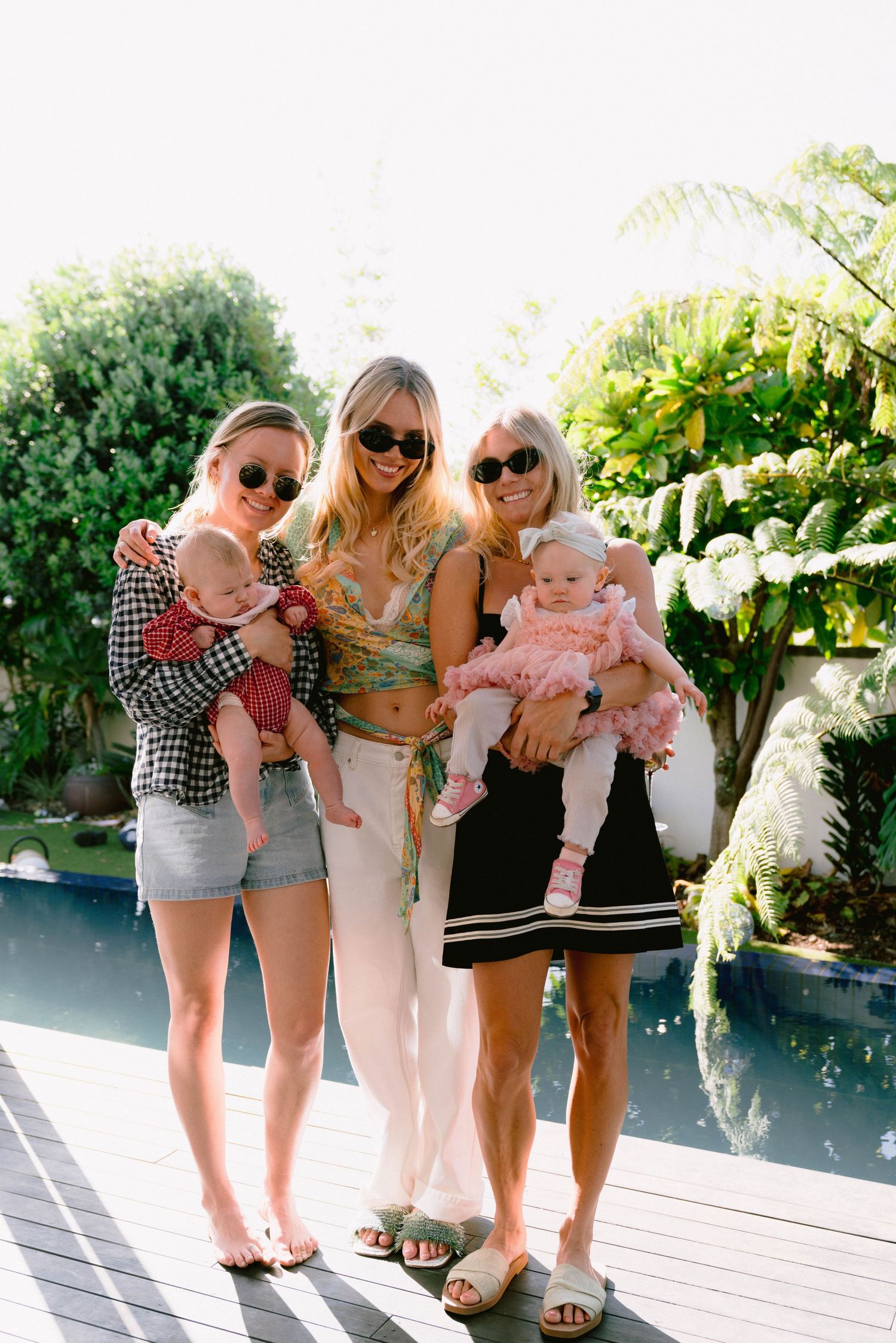 Three adults holding babies pose together on a sunny deck by pool with greenery.
