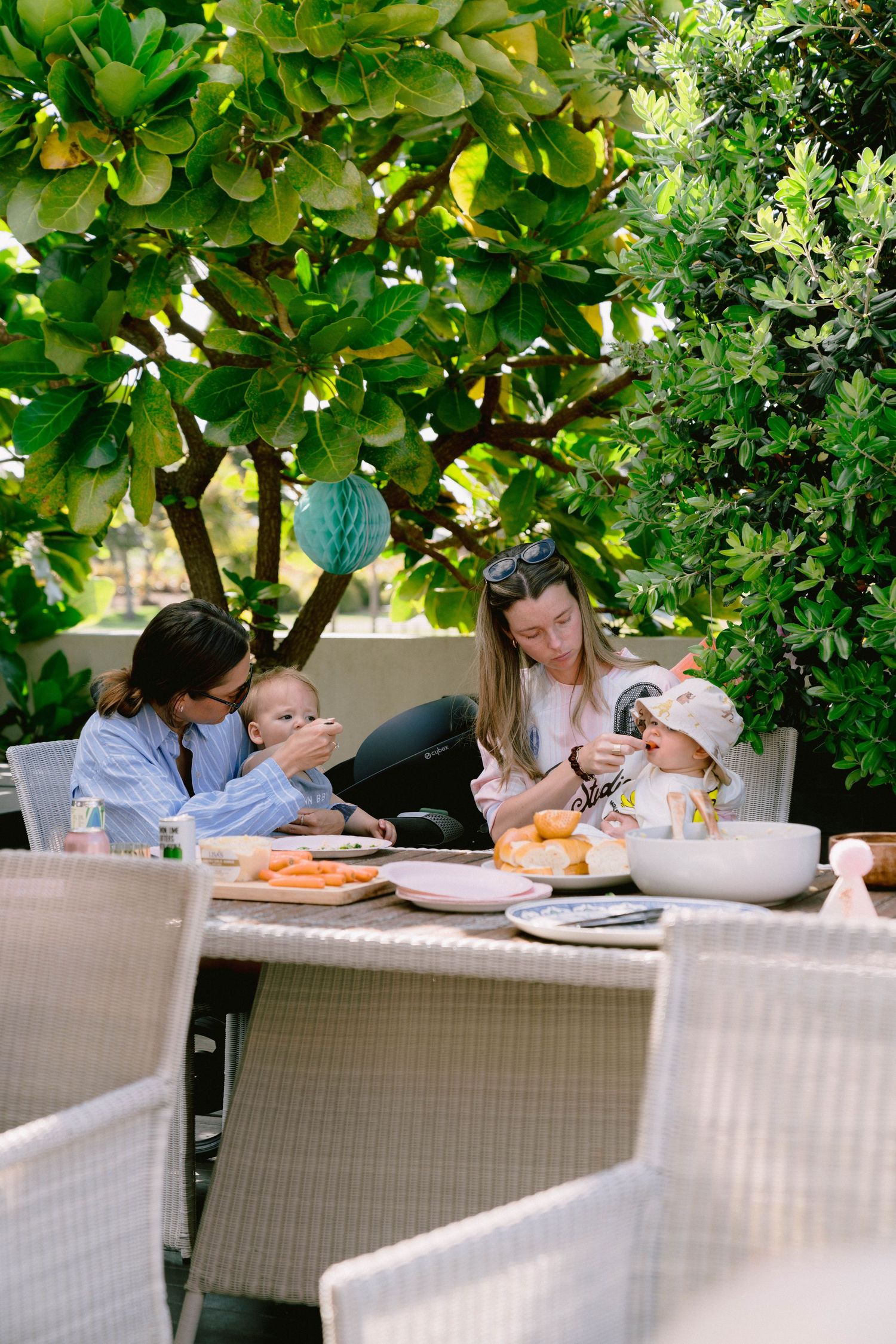 Guests seated at white outdoor furniture enjoying a meal under leafy trees.