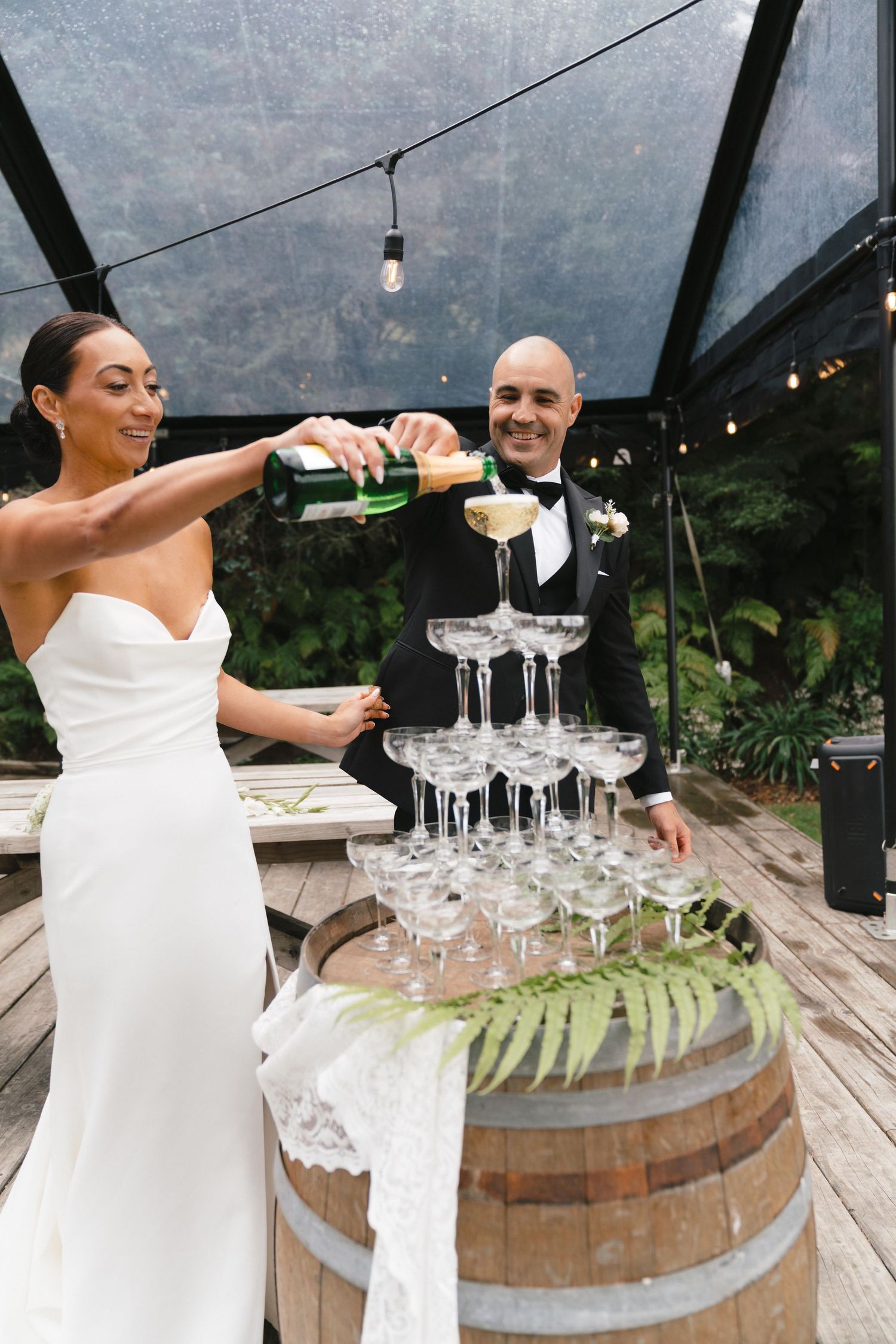 Wedding couple pours champagne into a tower of glasses arranged on a rustic wine barrel at an outdoor reception.