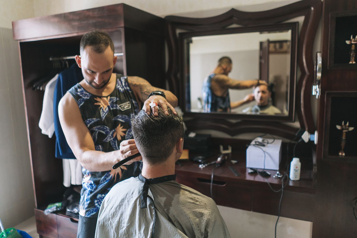 A barber wearing a patterned shirt gives a haircut to a client in a traditional barbershop setting with mirrors.