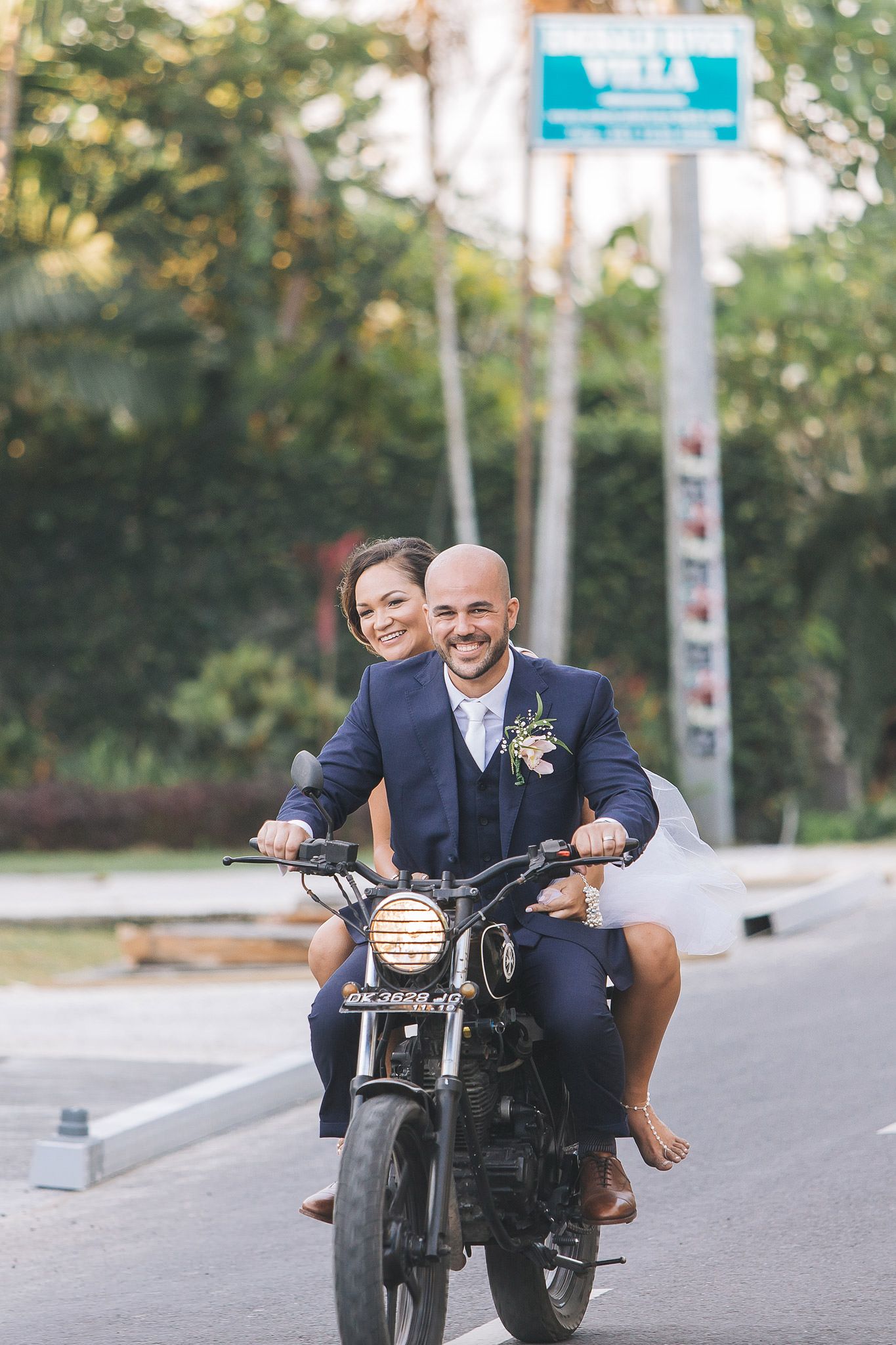 Wedding couple poses playfully on vintage motorcycle on urban street for creative photography session.