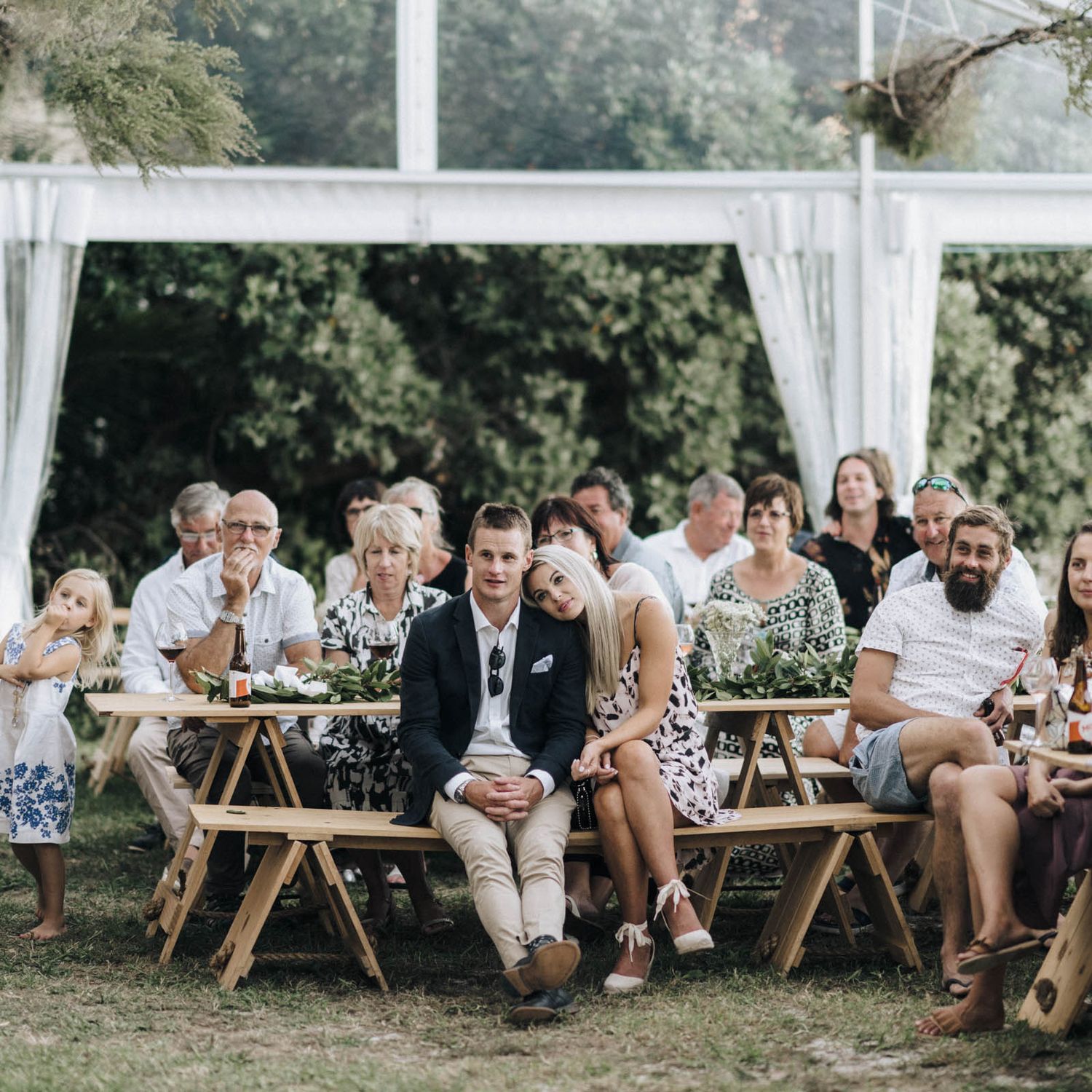 Wedding guests relax on wooden benches under an outdoor tent canopy during a summer celebration.