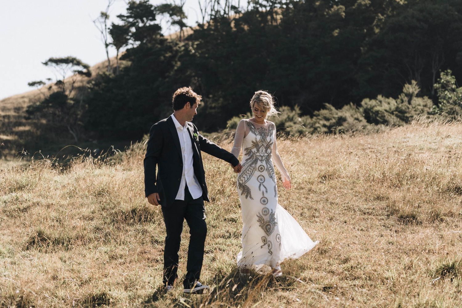 A couple in wedding attire holds hands while walking through a golden field on a sunny afternoon.