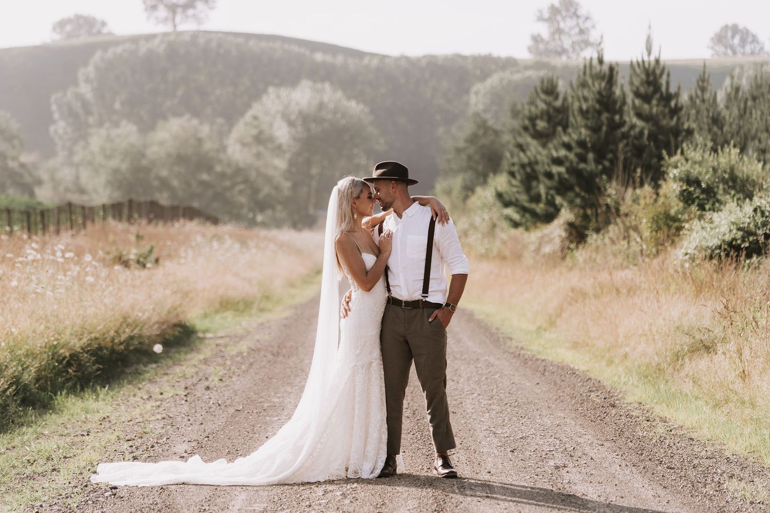 Newlyweds share romantic moments together on a rustic dirt path surrounded by misty golden fields at sunset.