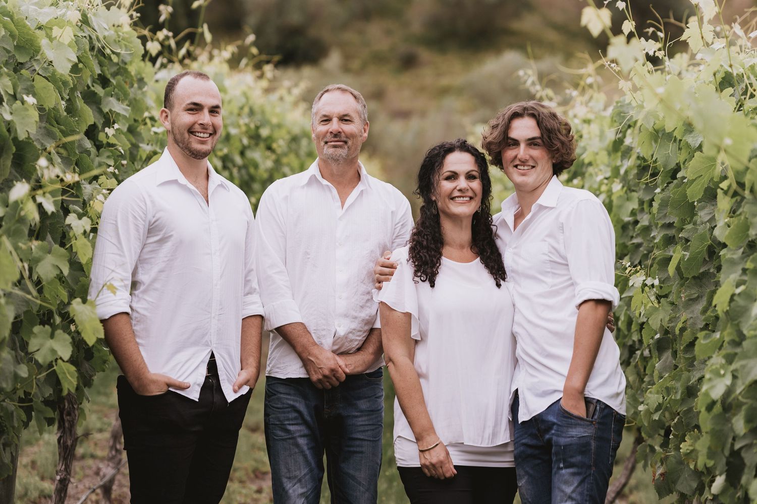 Four people in white shirts standing together between rows of green grapevines in a sunny vineyard.