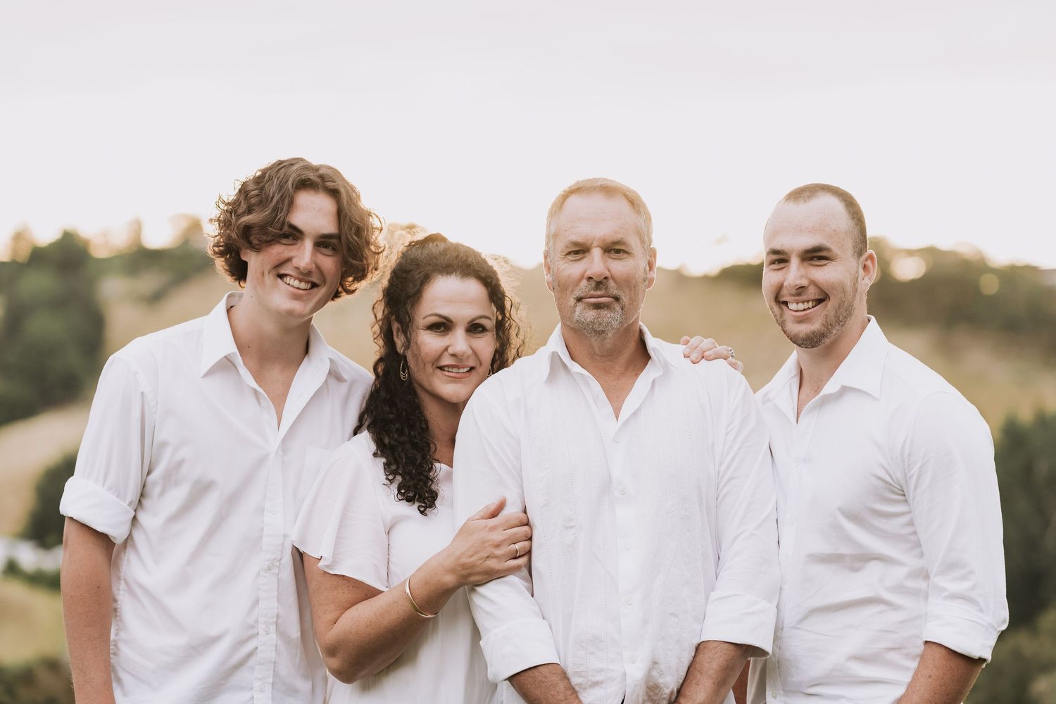 Group of four wearing white shirts posing together outdoors against a sunlit hillside background.