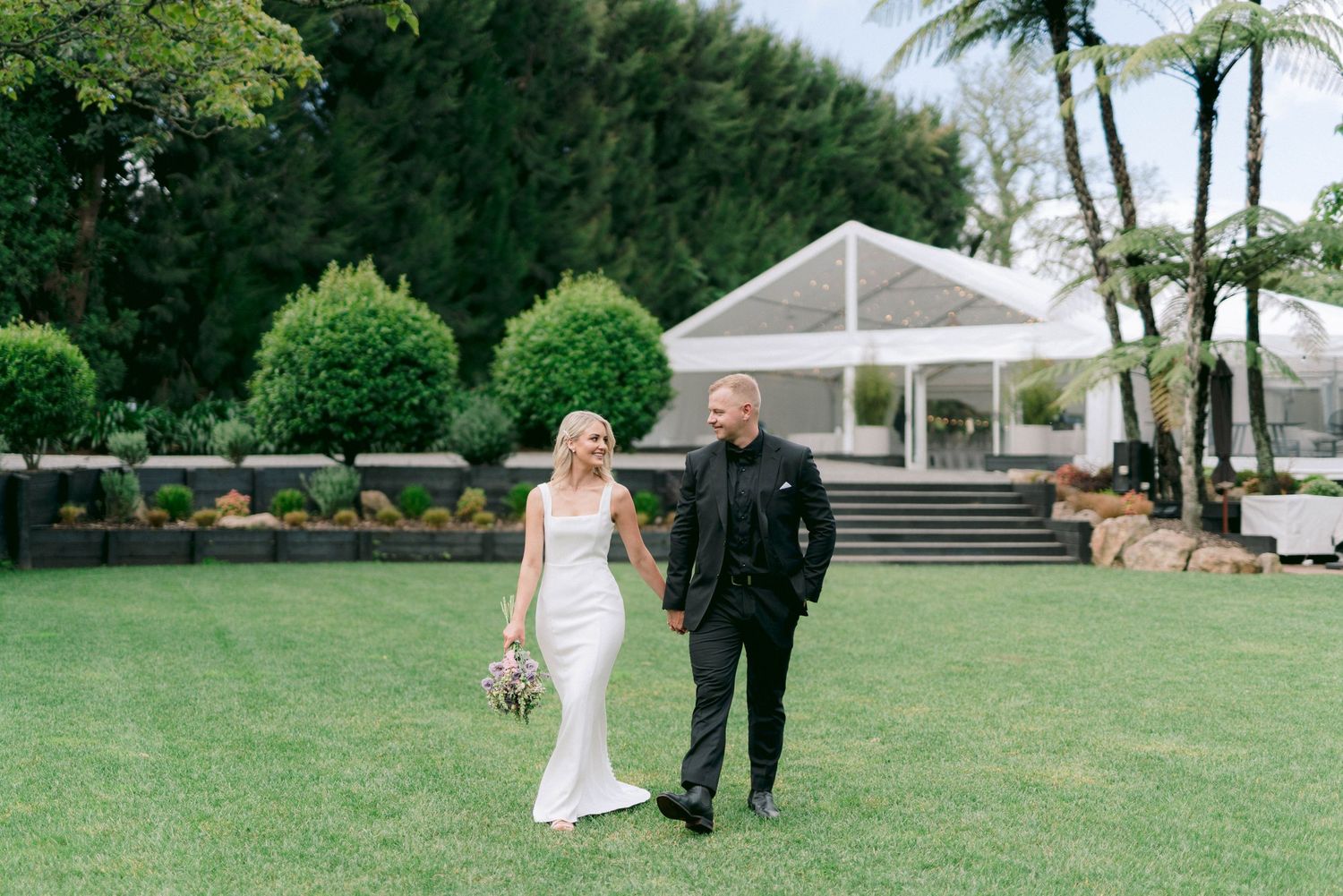 A couple walks across a manicured lawn with a white gazebo and palm trees in the background.