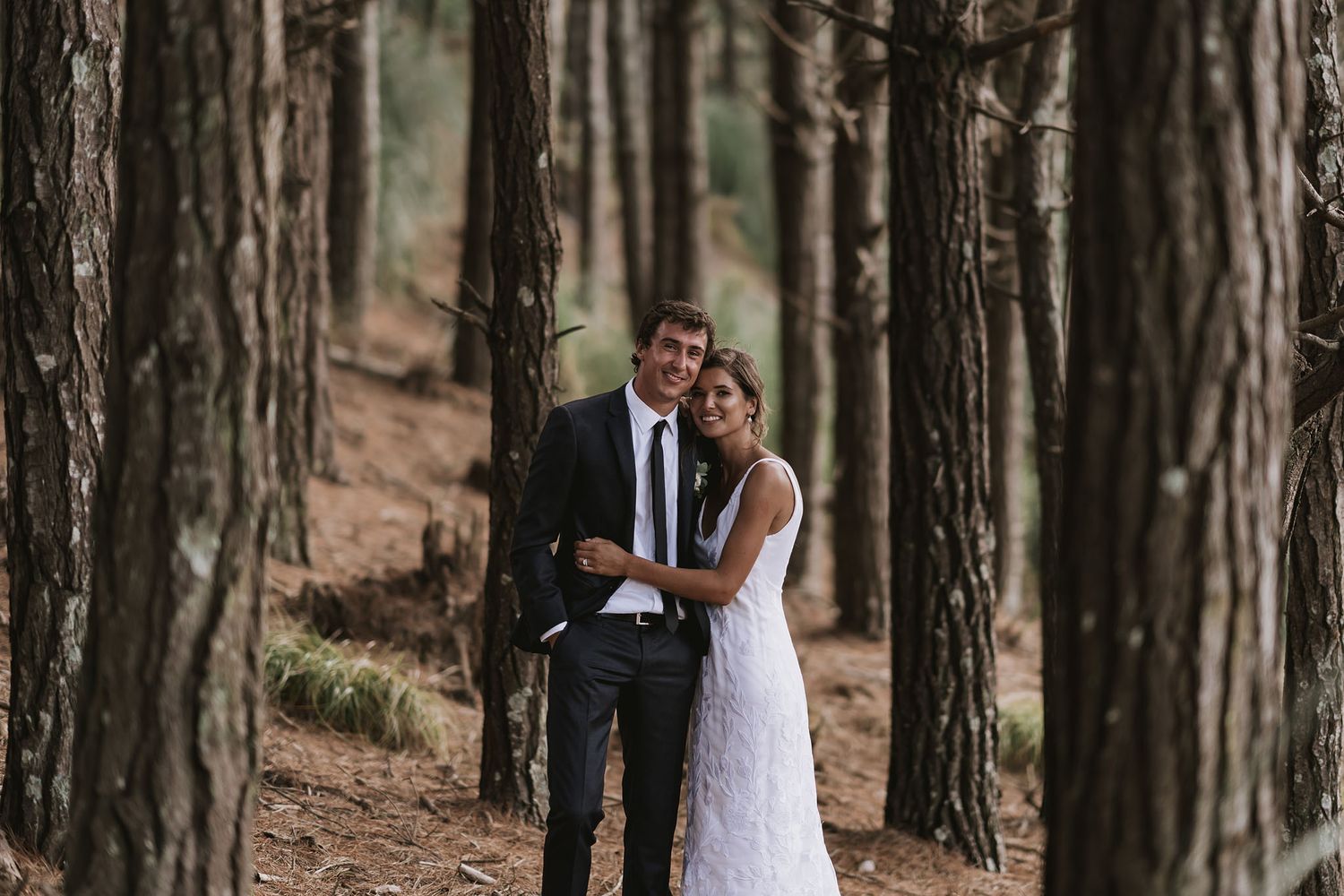 Couple in formal wedding attire embracing while standing in a serene forest of tall pine trees.