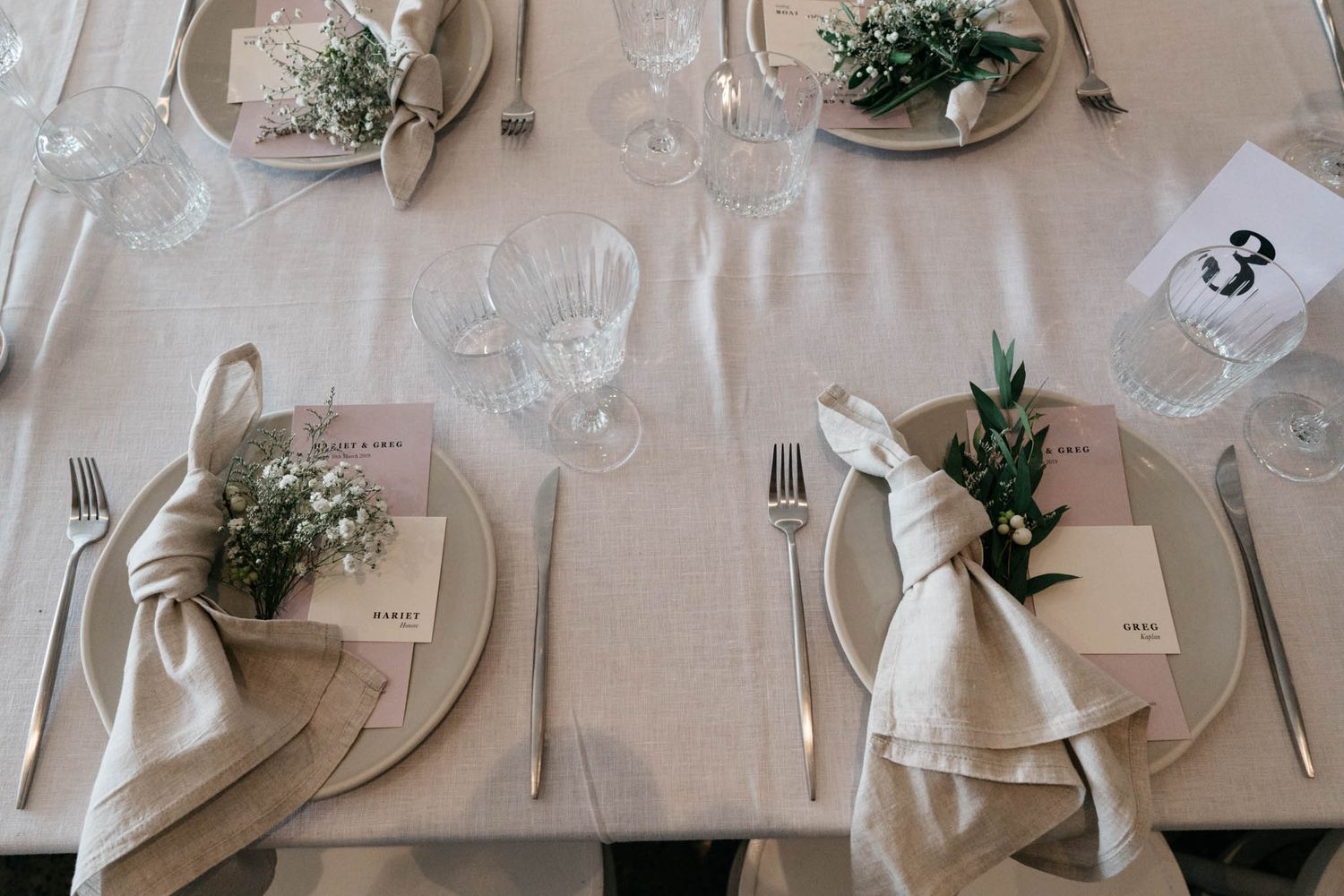 Elegant table setting with neutral linens, place cards, greenery sprigs, and wine glasses on a cream tablecloth.