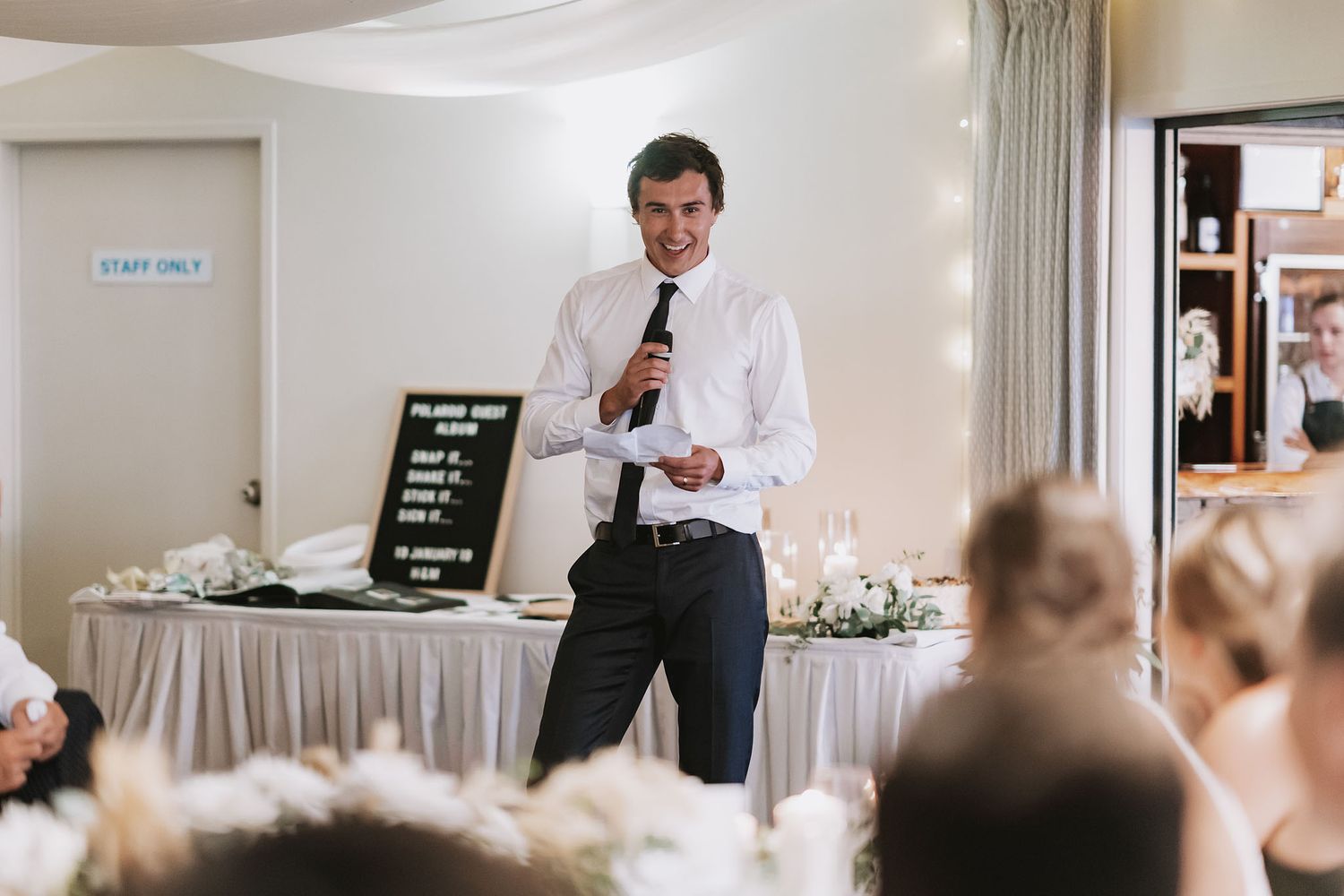 A person in formal attire delivers a speech at a wedding reception with tables and decor visible in the background.