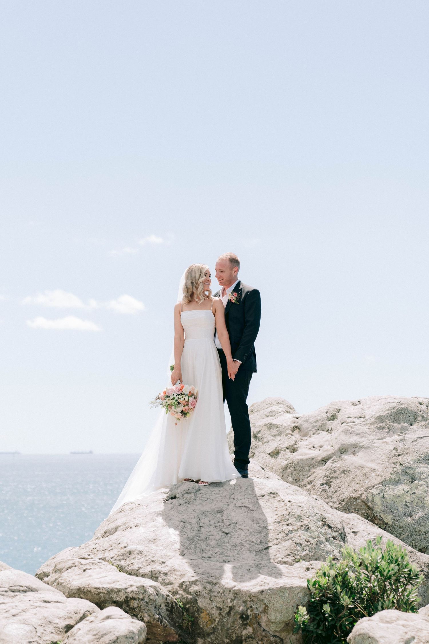 Newlywed couple stands embracing on coastal cliff with ocean view and bouquet on beautiful wedding day.