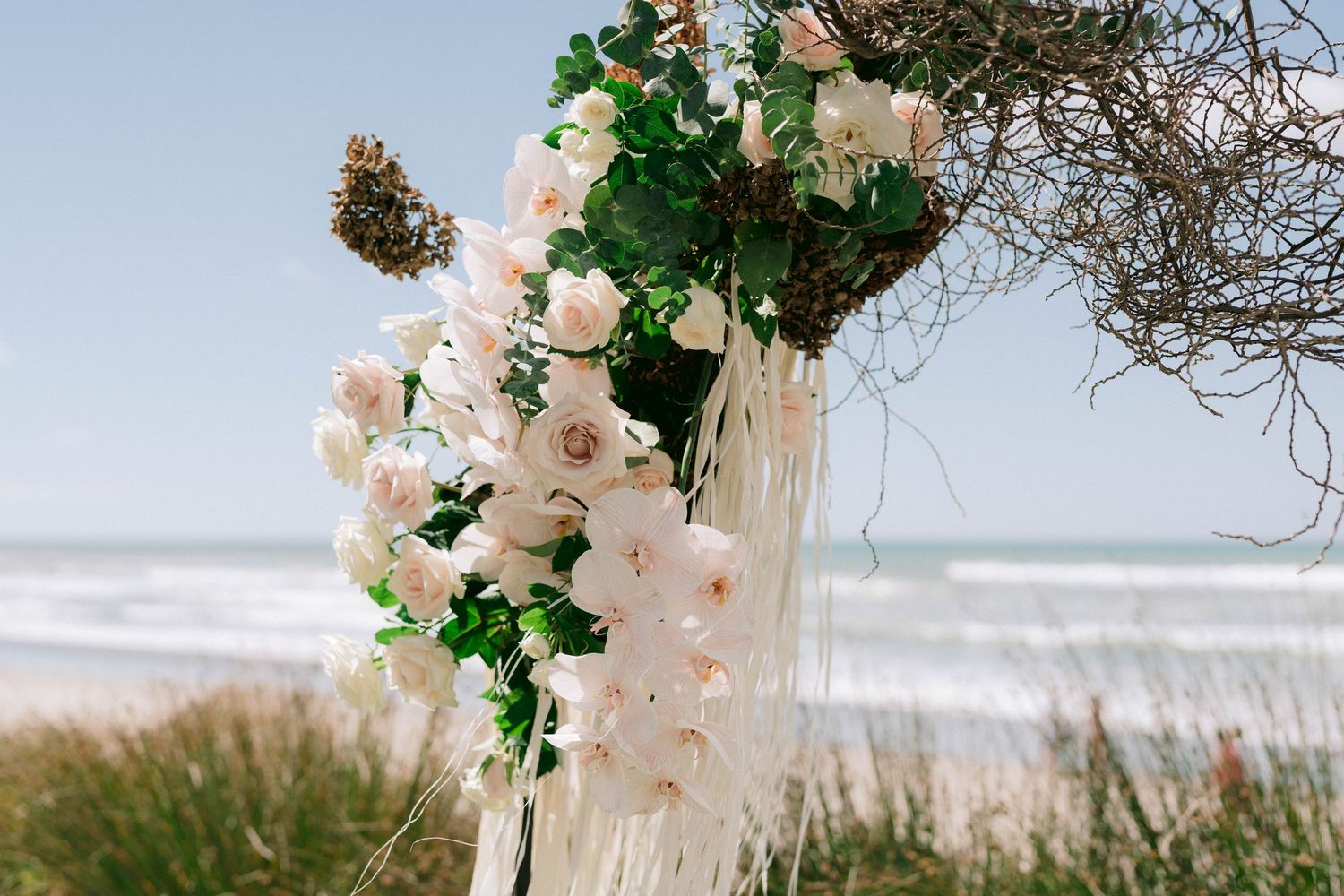 Delicate pink roses and greenery arranged on a wooden arch with ocean waves in the background.