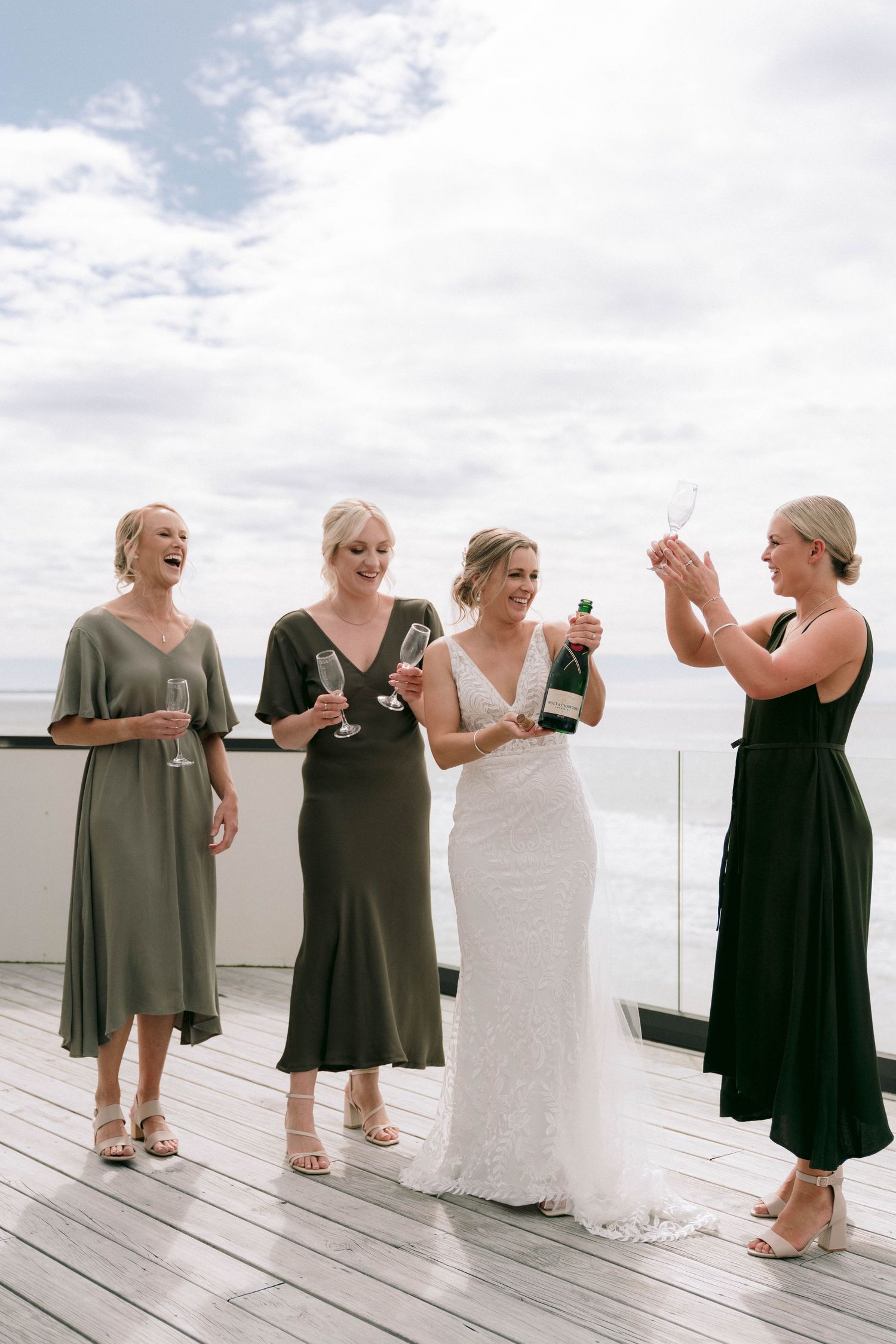 Group of bridesmaids in grey dresses laughing together on oceanfront deck.