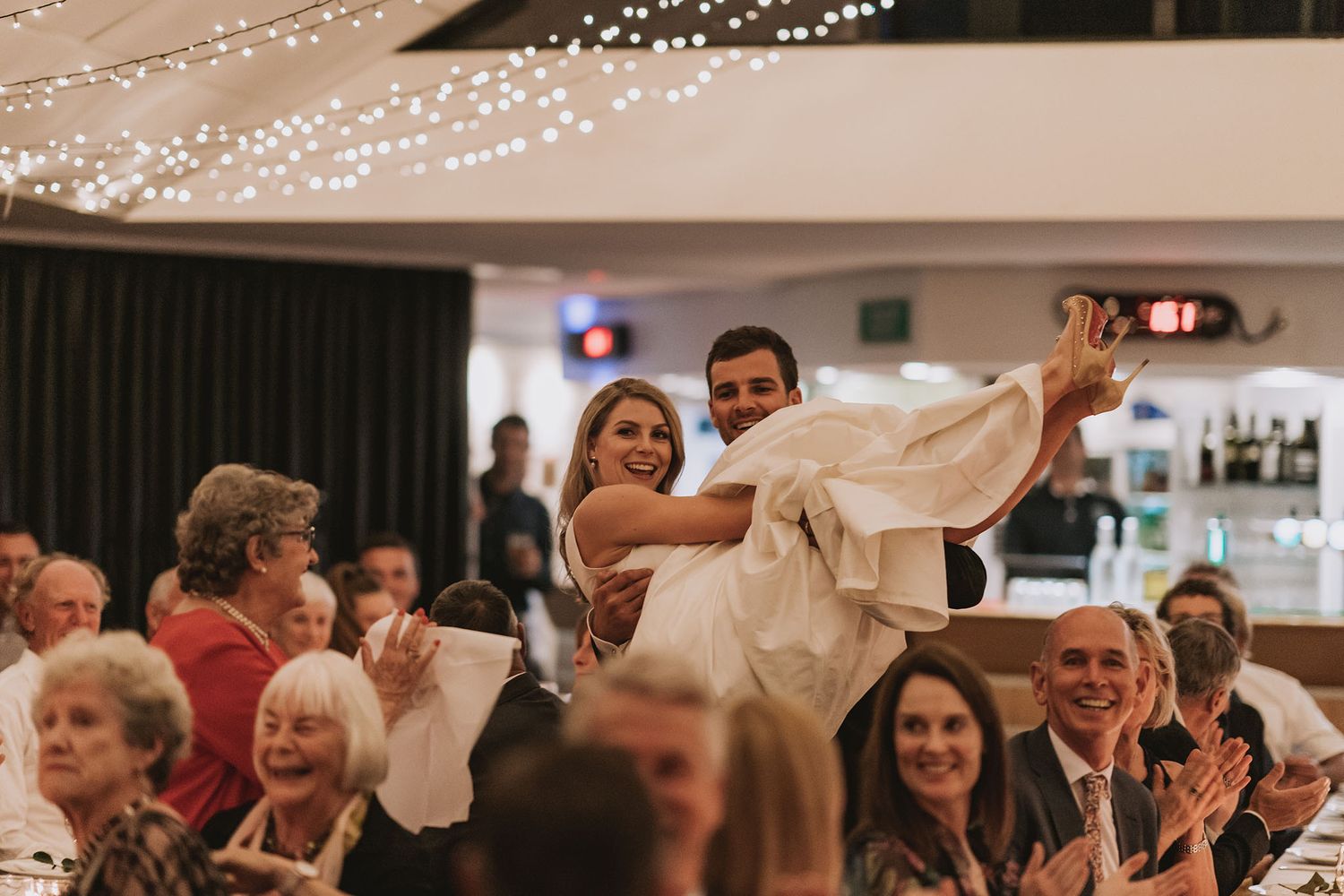 Wedding guests smile and applaud as couple performs a romantic dance lift during reception celebration.