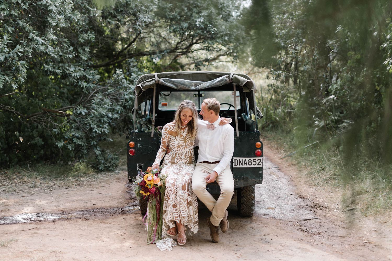 Romance and adventure meet as a couple sits on the back of a safari vehicle on a dirt road surrounded by lush greenery.