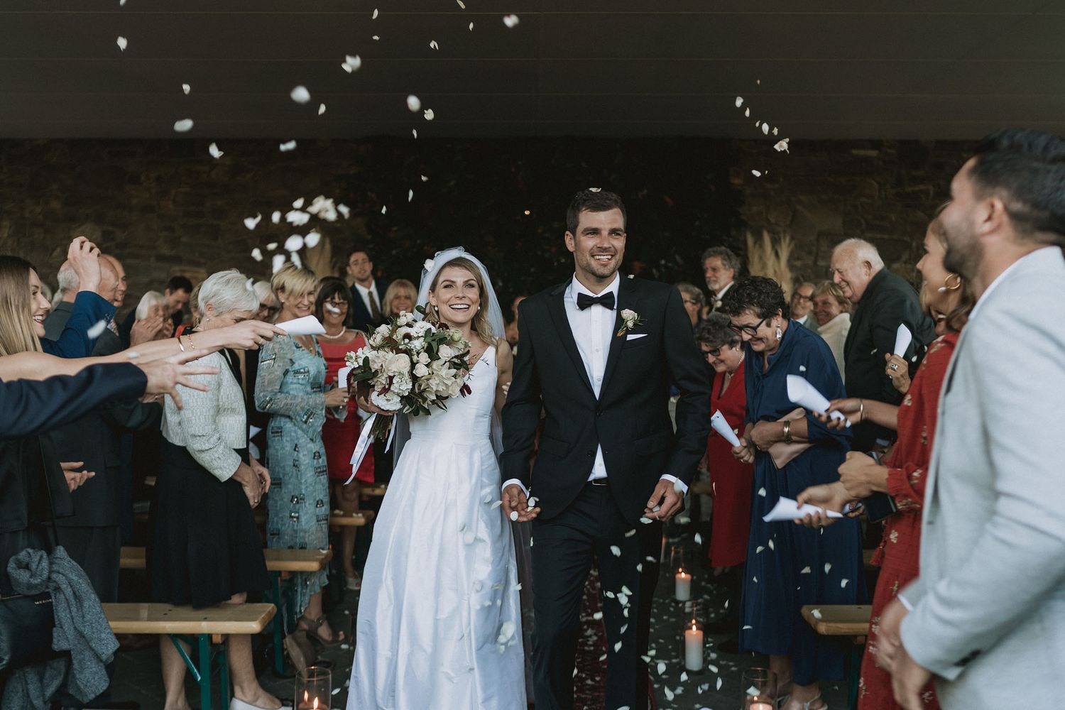 Wedding guests toss confetti over the celebrating couple as they walk under a shower of white petals during reception.