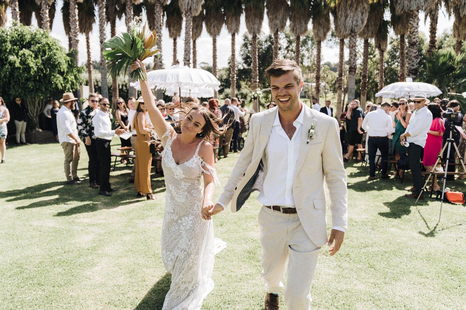 Newlyweds walking hand in hand on lawn while wedding guests celebrate.