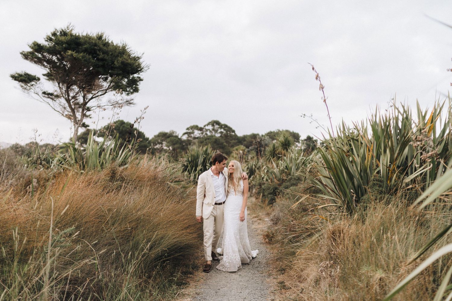 Couple in white walking down a coastal path with native plants and trees in the background.