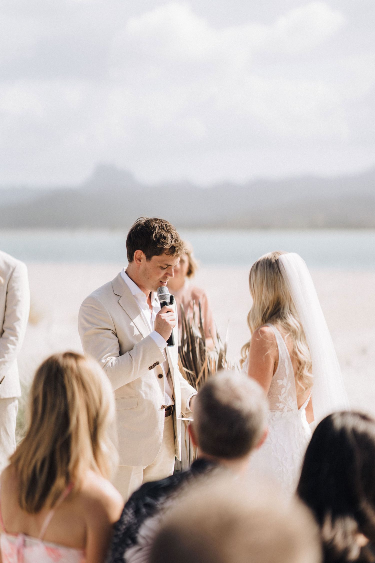 Guests gather at an elegant seaside wedding ceremony with white and cream decor under cloudy skies.