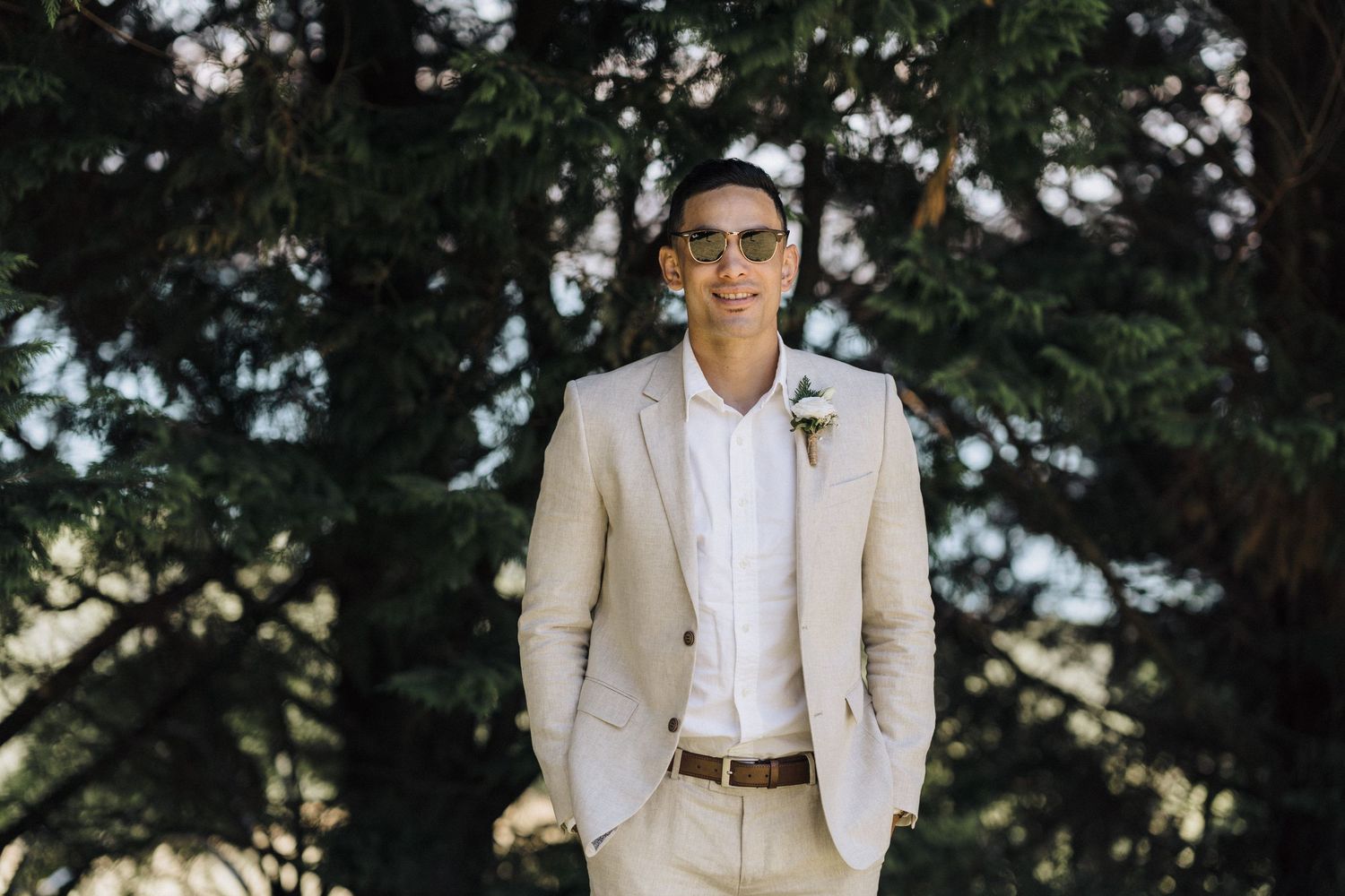Groom in a light beige suit and white shirt stands outdoors in a romantic garden setting under natural lighting.