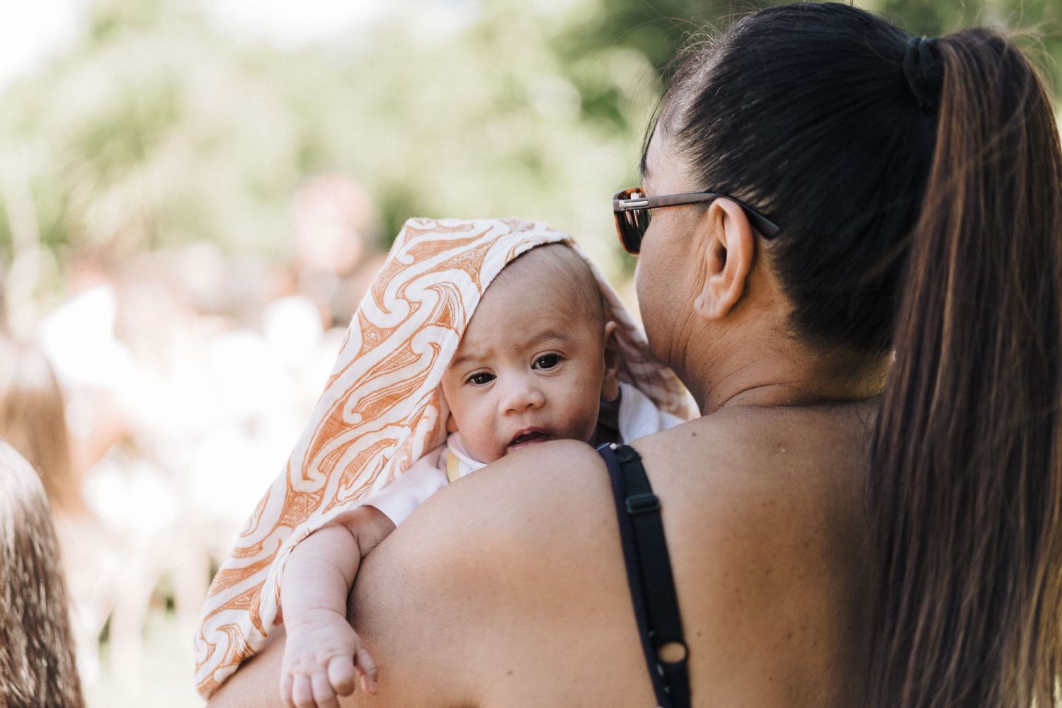 Mother holding baby with pink headband outdoors in natural lighting.