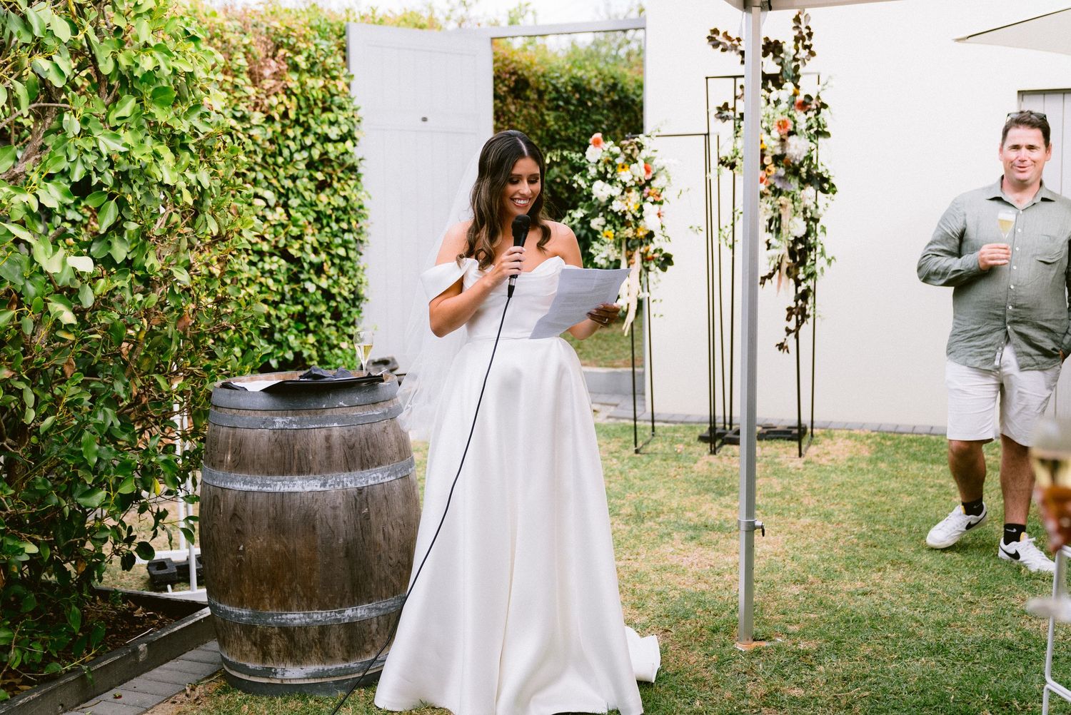 A bride reads from papers while standing next to a rustic wine barrel in a garden setting.