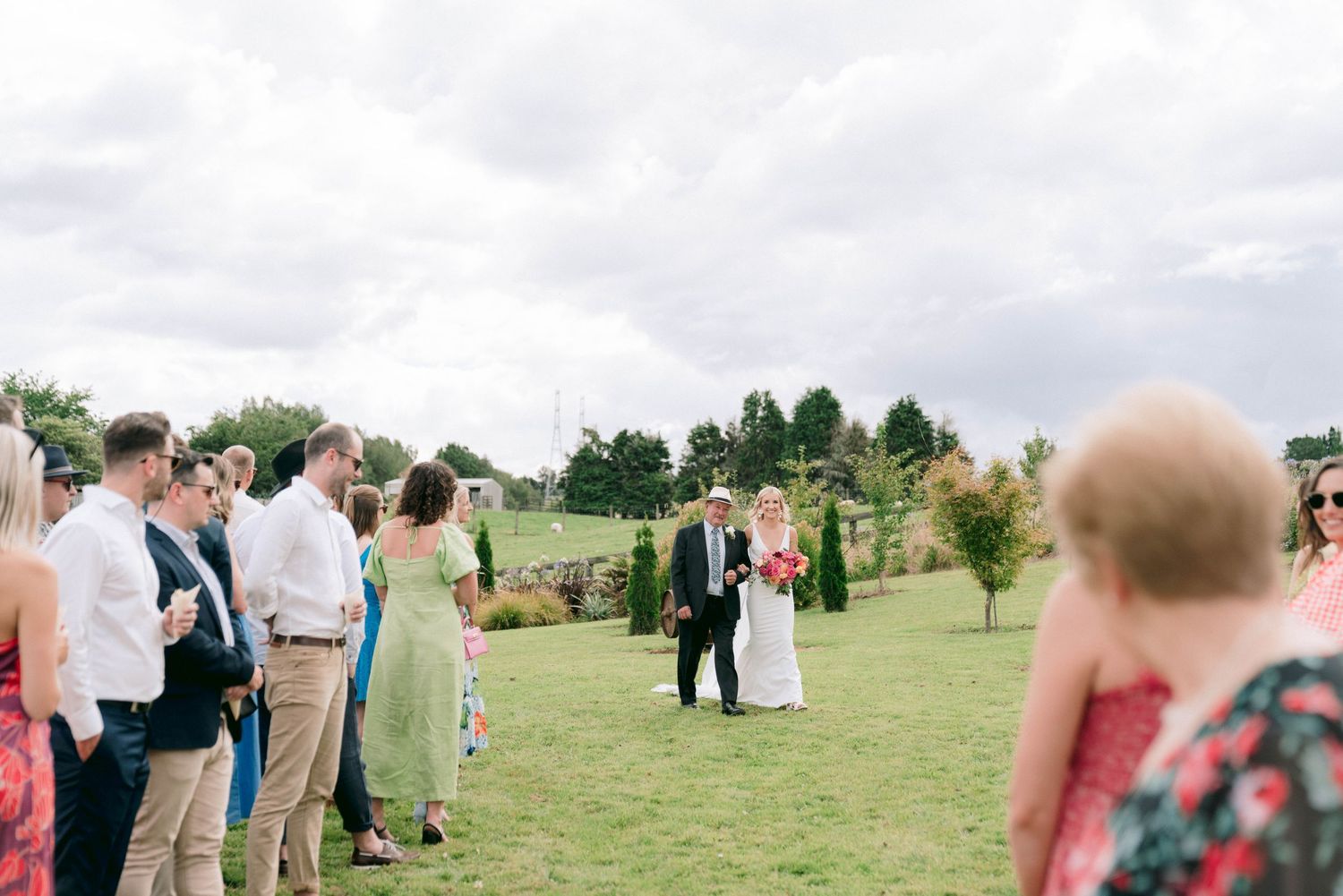 Wedding guests gather on lawn while ceremony takes place in background.
