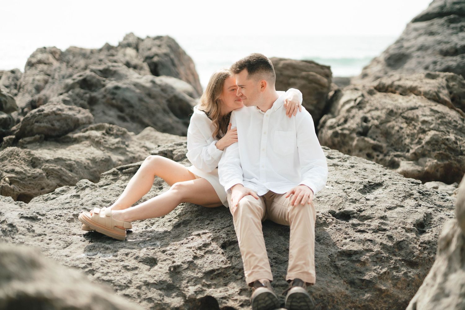 A couple sits intimately on coastal rocks during a romantic beach photoshoot at sunset.