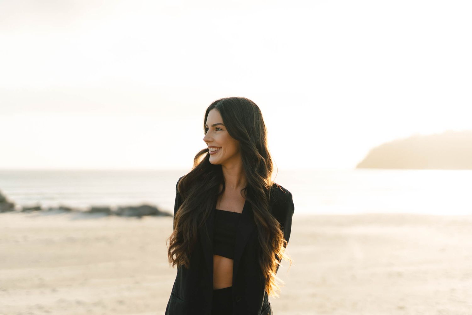 A person in black attire poses against a warm sunset backdrop near the ocean.