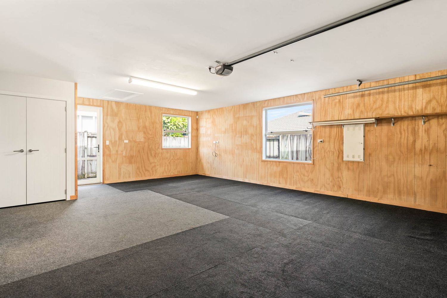 Empty garage interior with plywood walls, gray carpet flooring, and fluorescent lighting.