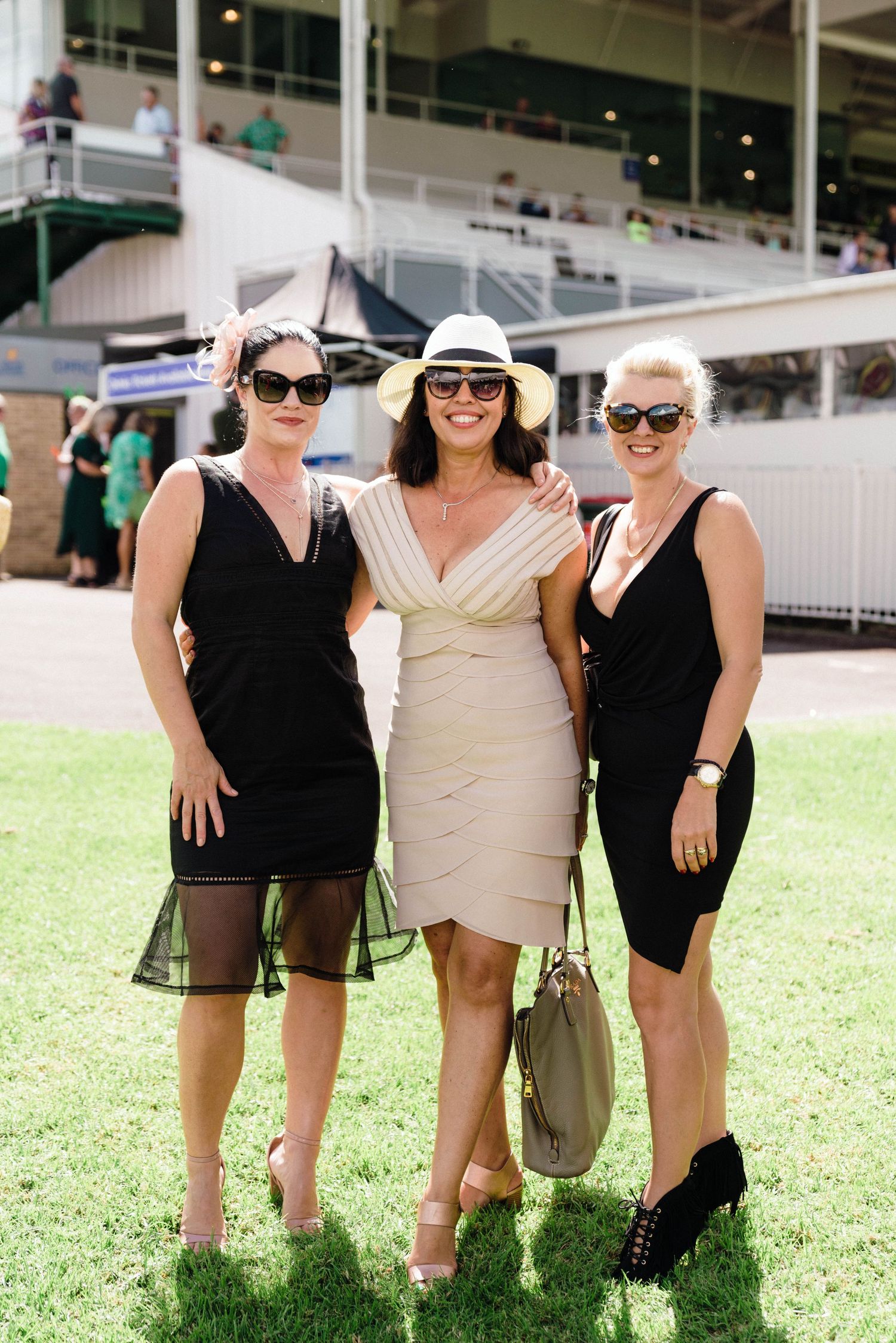 Three friends wearing summer outfits pose together on a grassy lawn area at an outdoor event.