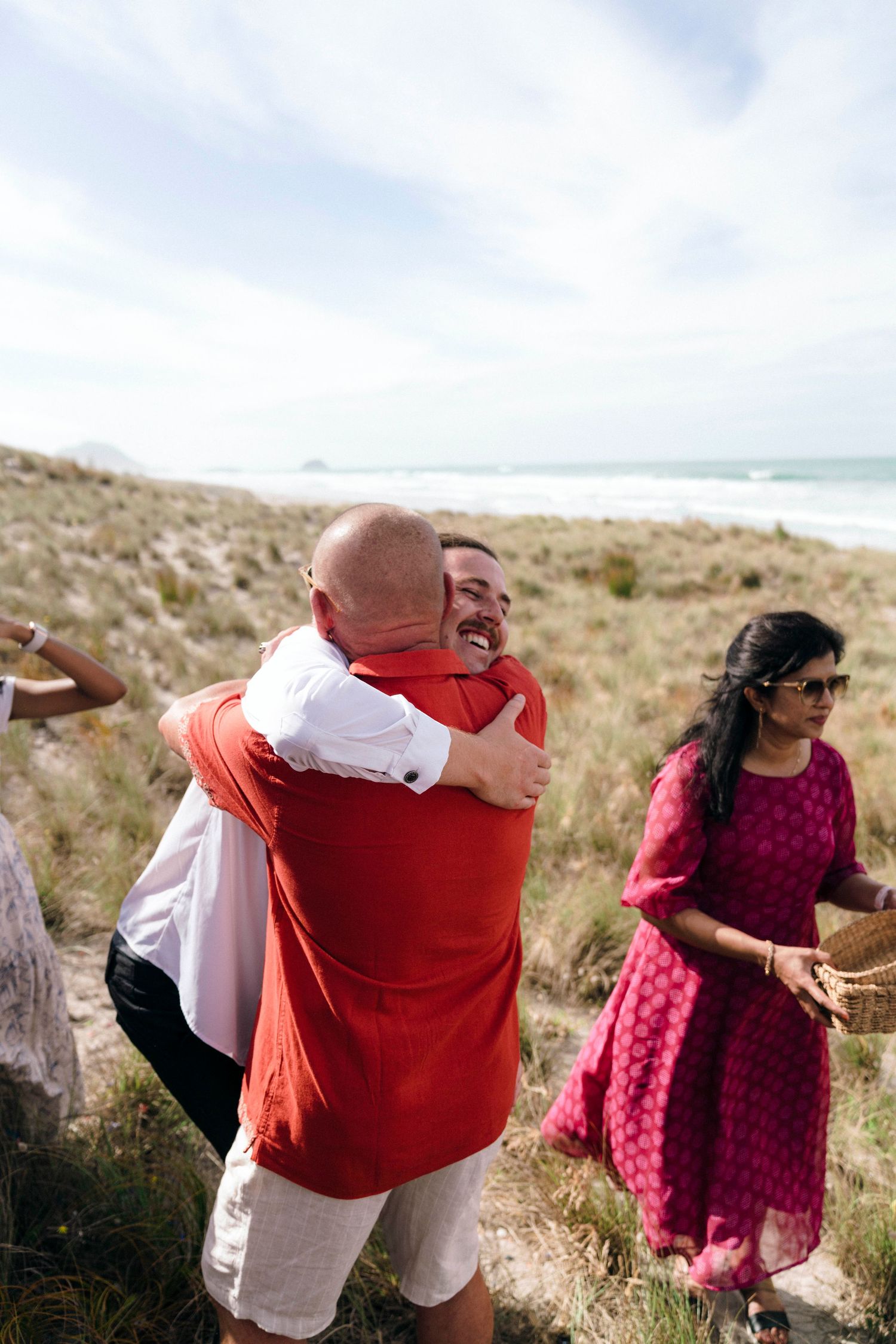 Happy guests in white and coral outfits embrace on a coastal hillside during a wedding celebration.