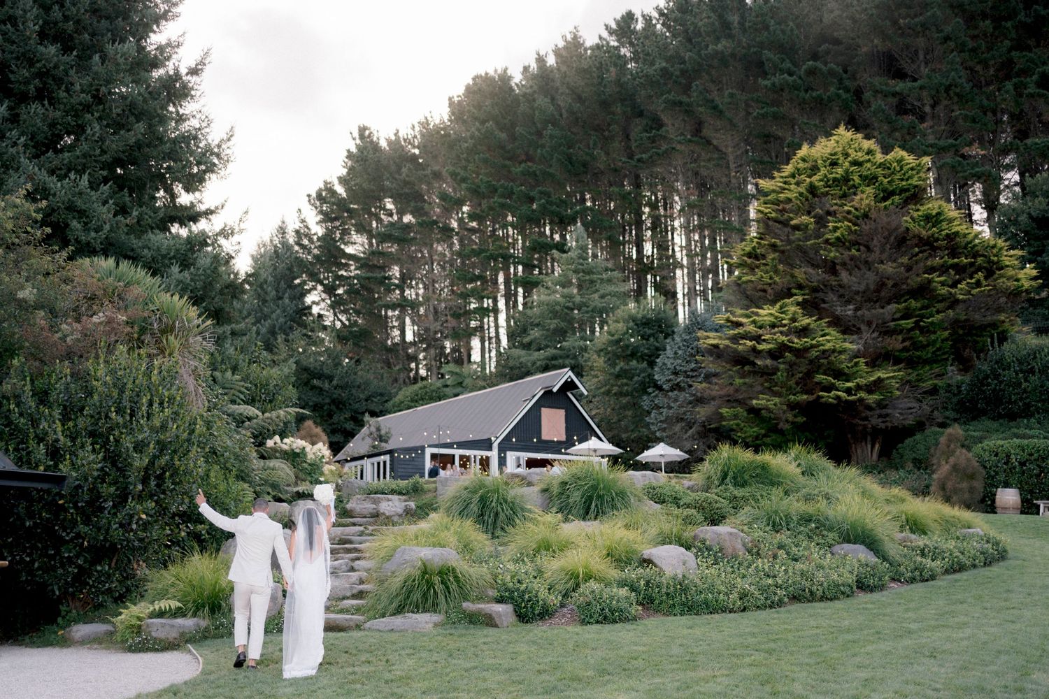 Wedding couple walking down landscaped path towards charming cottage in pine forest.