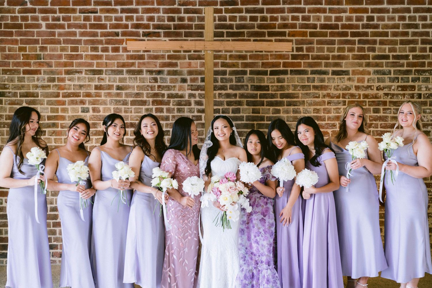 Bridesmaids in matching lavender dresses stand together against a brick wall holding bouquets at a wedding celebration.