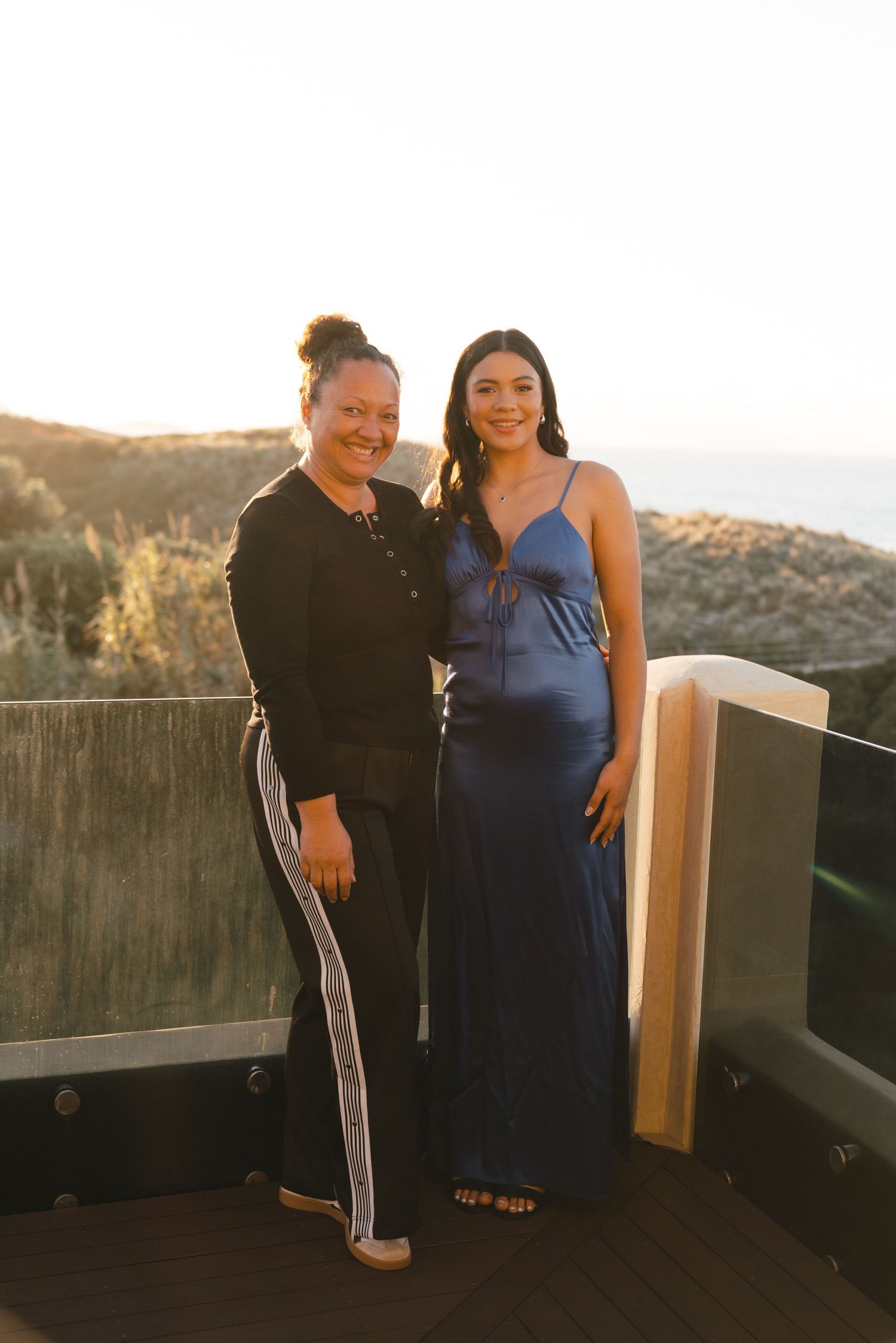 Two friends posing together on balcony during golden hour wearing contrasting formal attire.