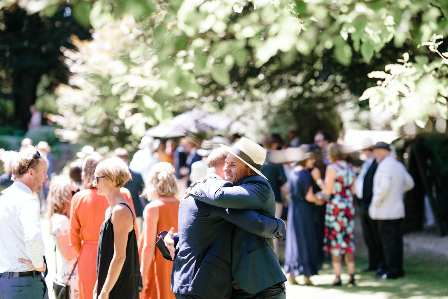 Wedding ceremony attendees stand together on a sunny day surrounded by lush greenery and trees.