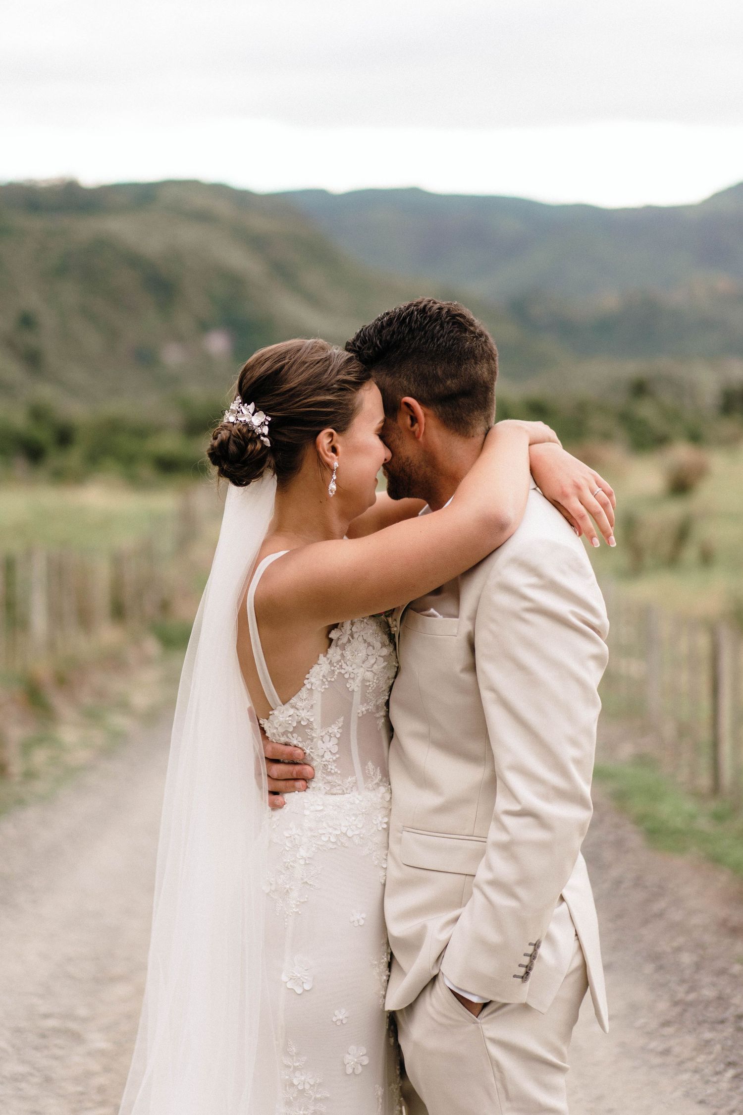 A romantic wedding moment on a countryside dirt road with mountains and fences in the background.
