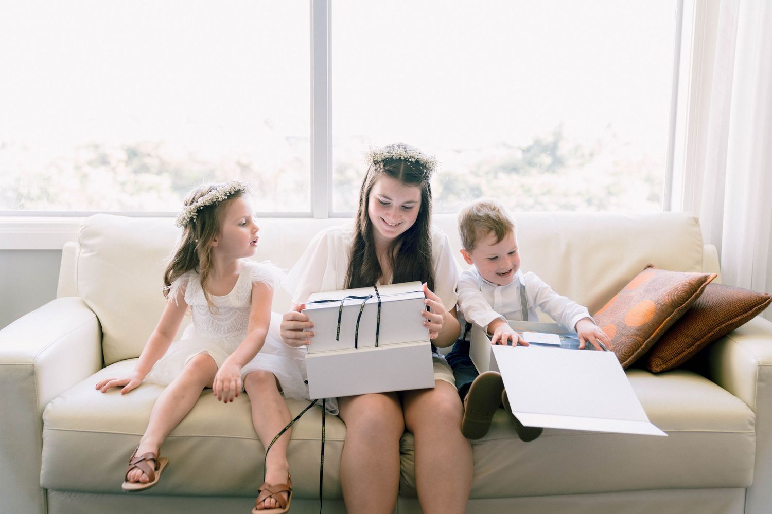 Three children sit together on a white couch reading papers and documents in a bright sunlit room.