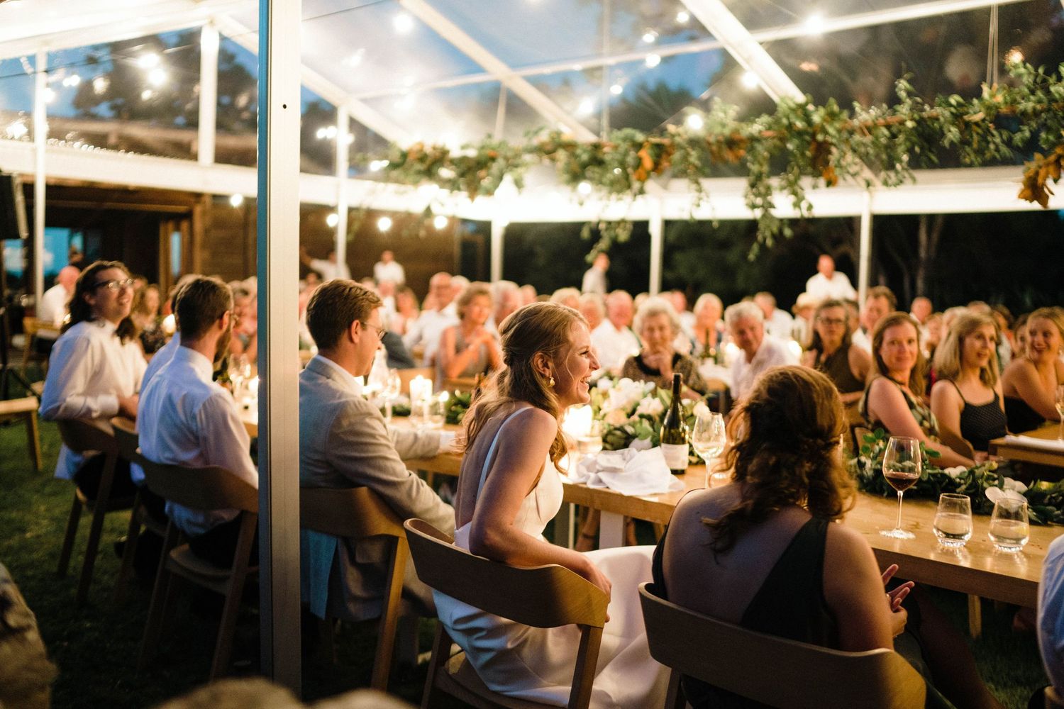 Wedding guests enjoy an outdoor reception under a white tent with string lights and greenery.